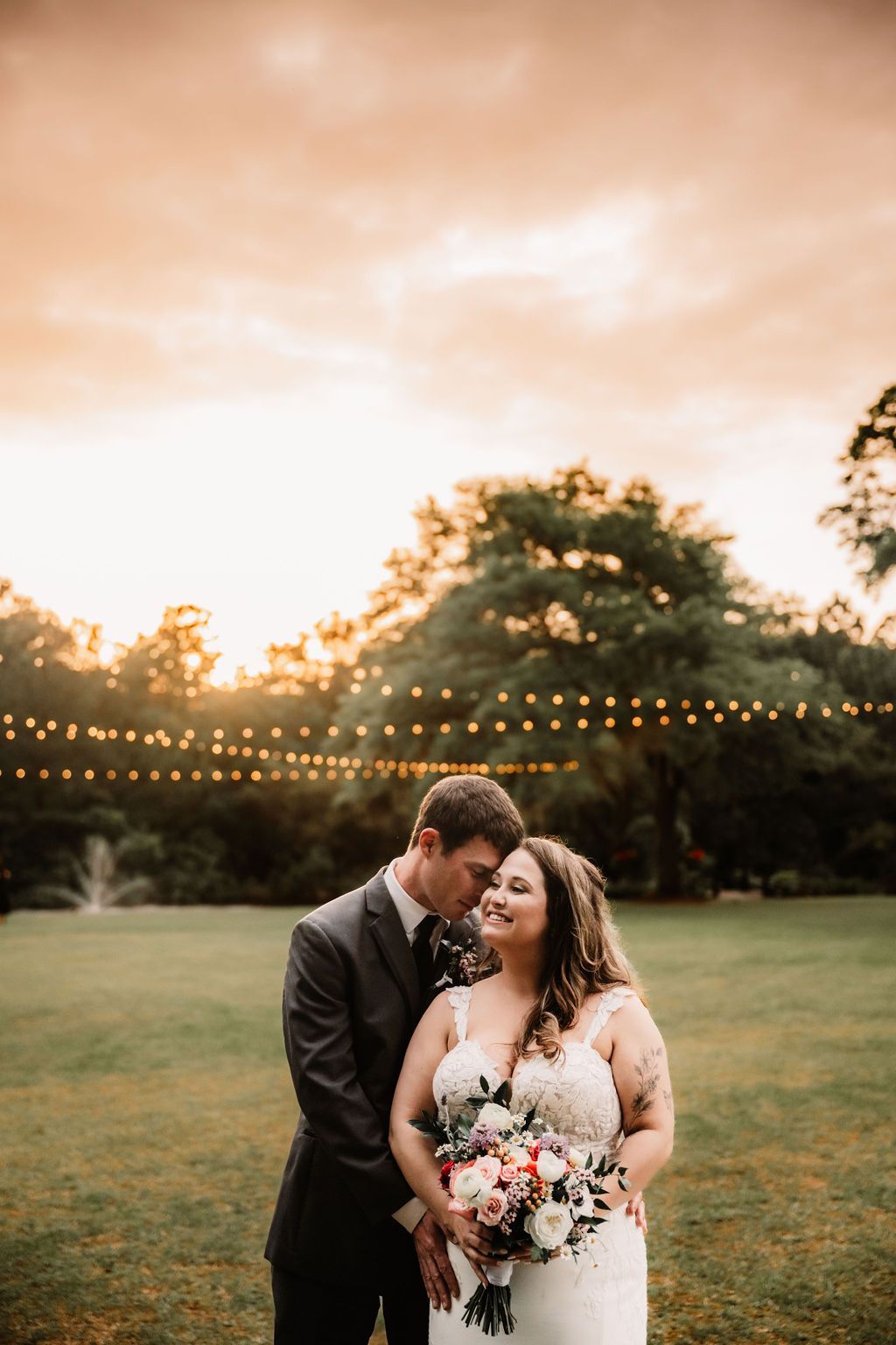 Bride and groom embracing in a field at sunset with string lights overhead