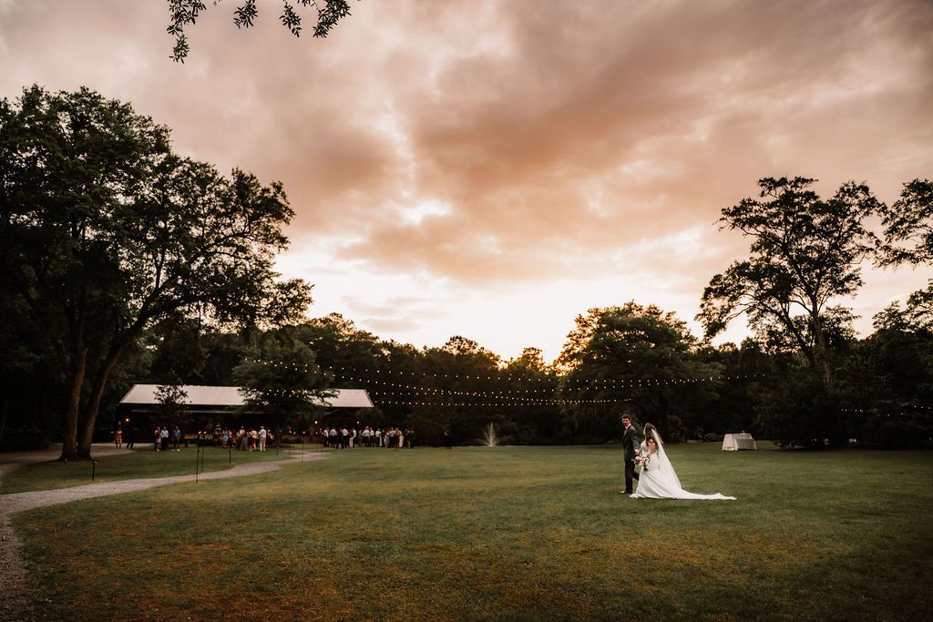 Bride and groom standing on a grassy field at sunset, framed by trees and warm sky