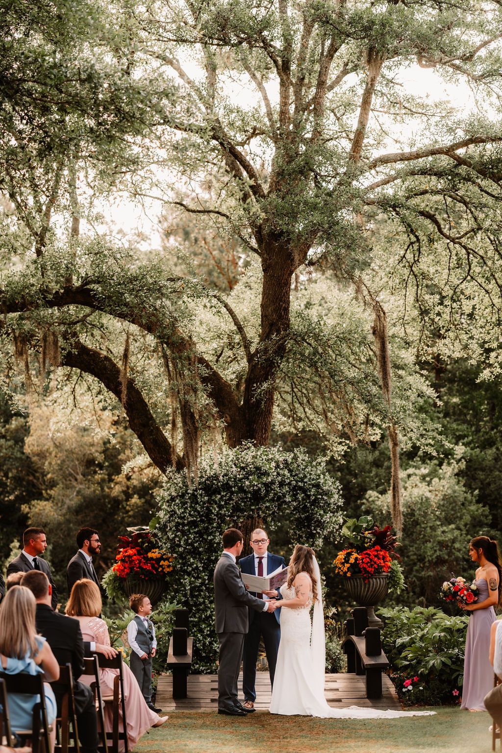 Outdoor wedding ceremony under oak trees, with couple standing at the altar surrounded by guests.