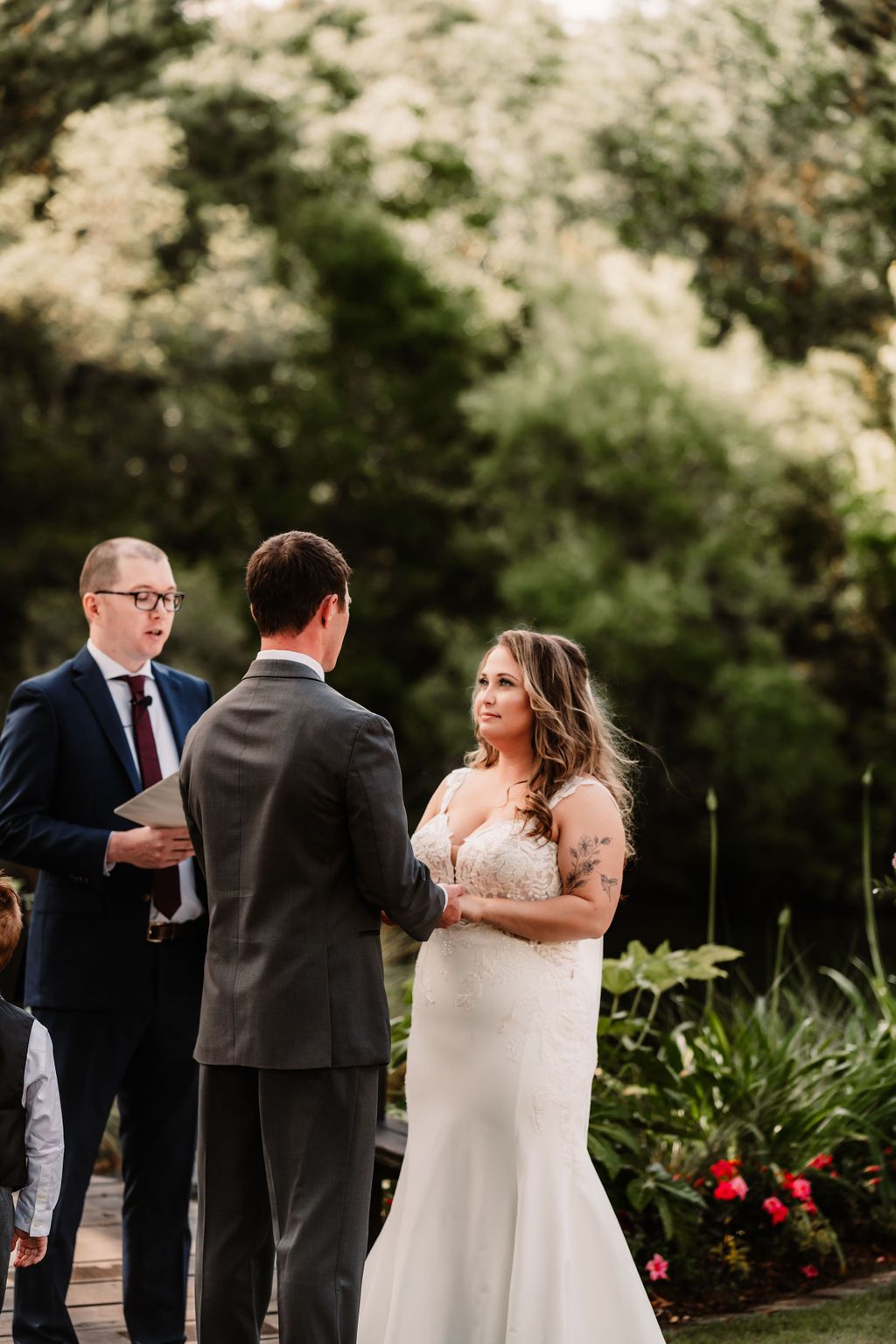 Wedding couple exchanging vows outdoors while an officiant stands nearby, with greenery and flowers in the background