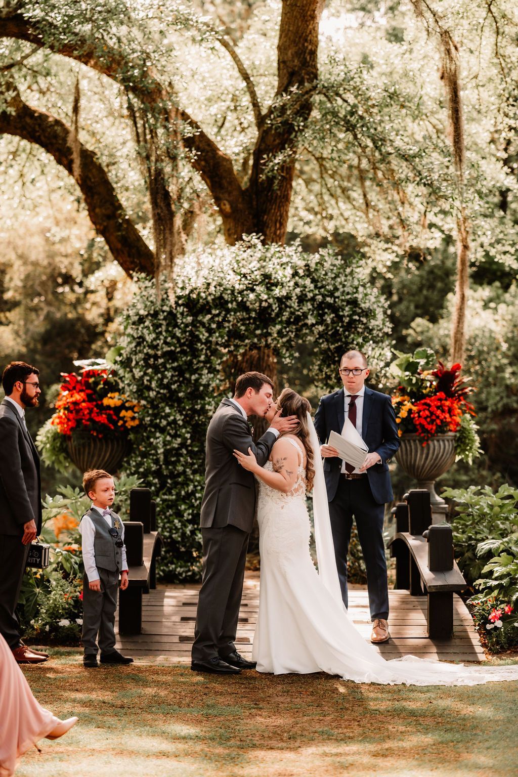 Outdoor wedding ceremony with bride and groom standing under trees, surrounded by floral arrangements and guests.
