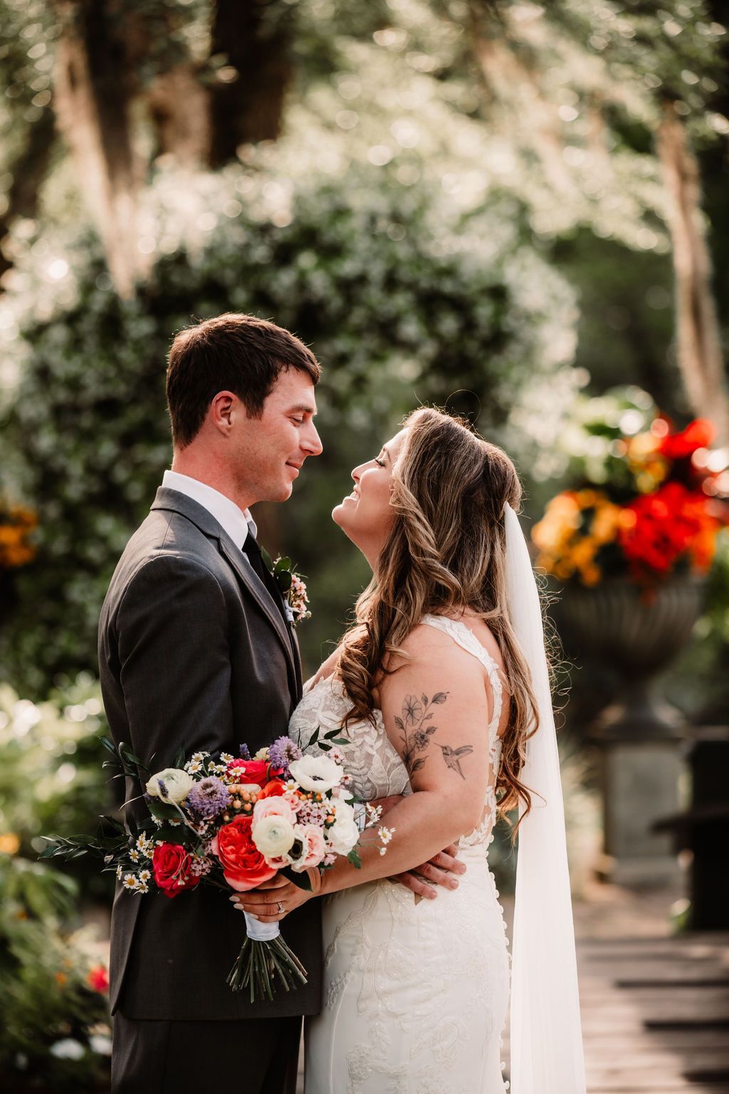 Bride and groom smiling in wedding attire outdoors, holding a bouquet amid colorful flowers.
