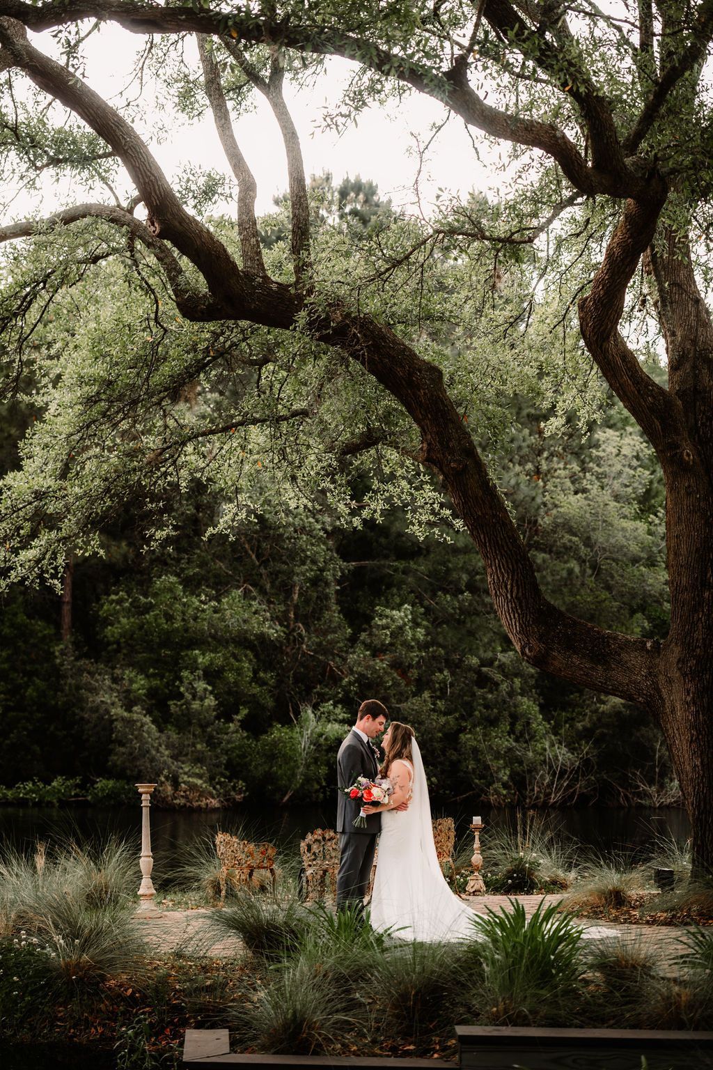 Bride and groom standing in a garden beneath large trees during an outdoor wedding ceremony