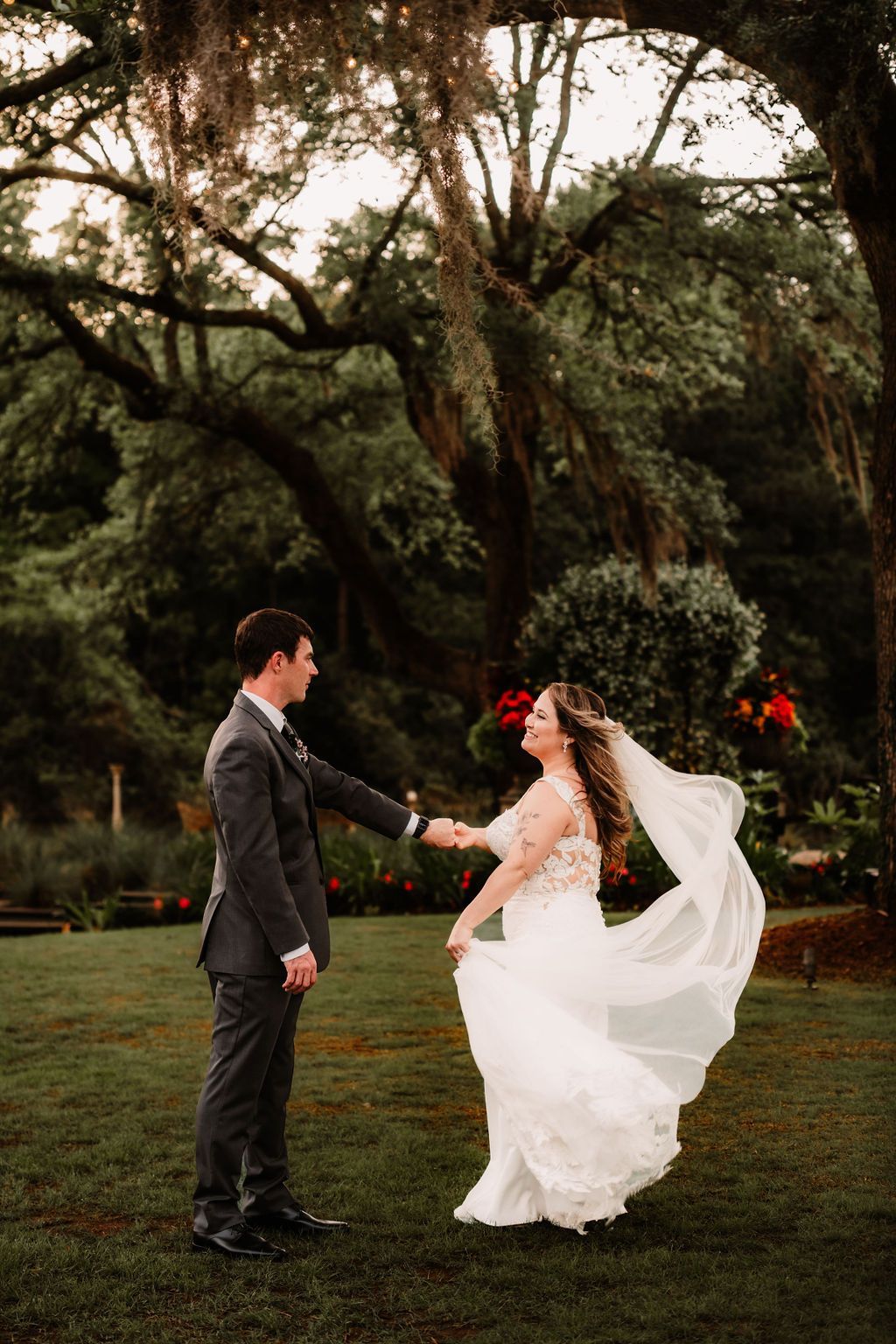 Couple in wedding attire holding hands in a garden under tall trees, bride’s veil flowing