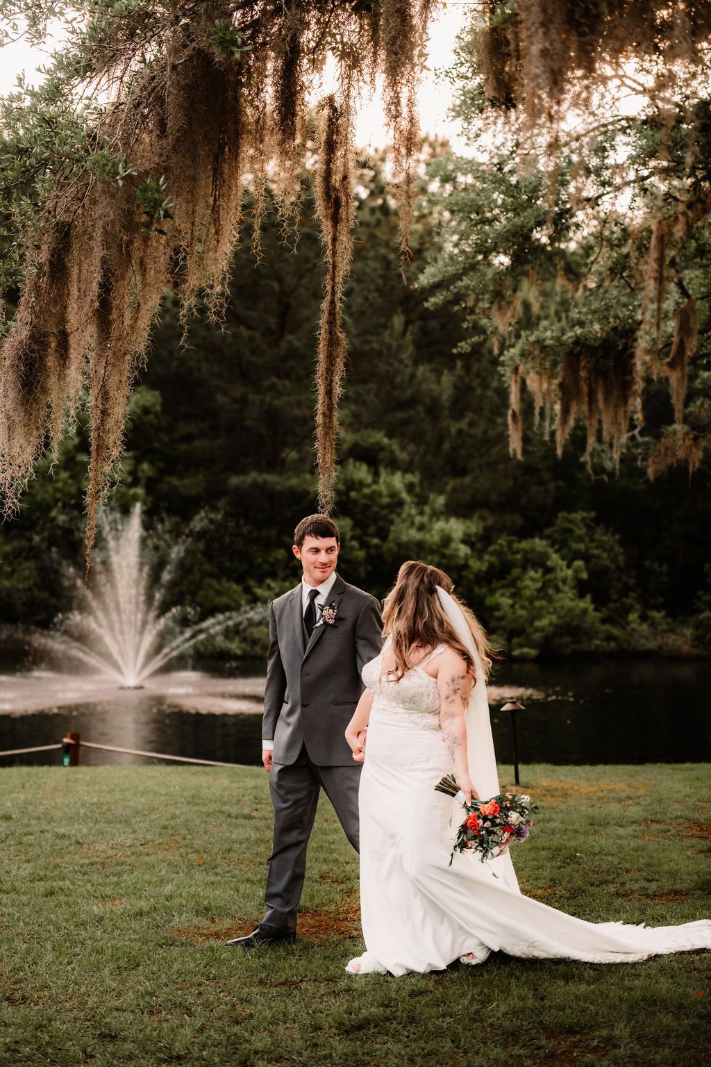 Bride and groom walking on grass beside a fountain in a garden with hanging moss trees