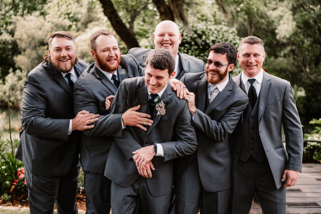 Group of six people in dark suits laughing together outdoors among trees.