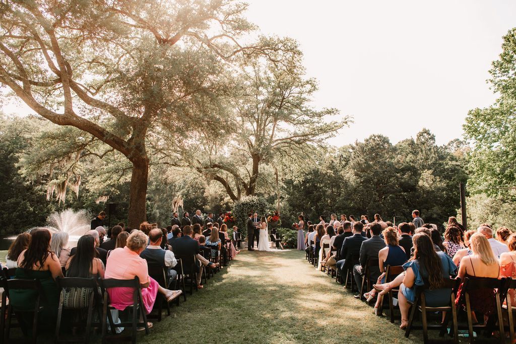 Outdoor wedding ceremony with guests seated on chairs under trees in a grassy garden