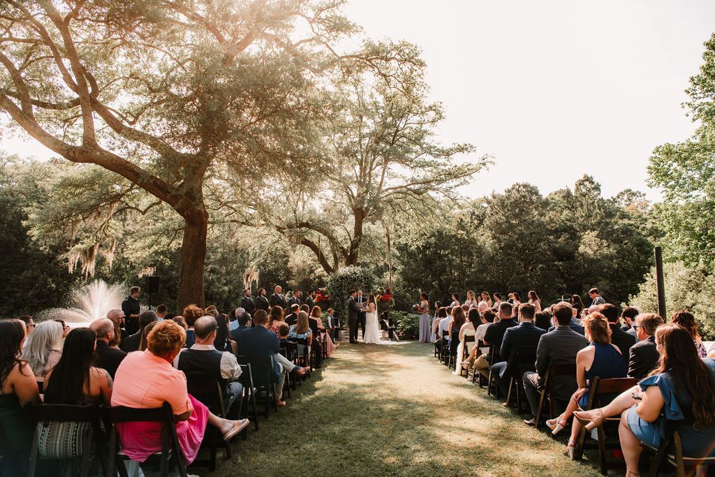 Outdoor wedding ceremony with seated guests under trees on a grassy lawn at sunset