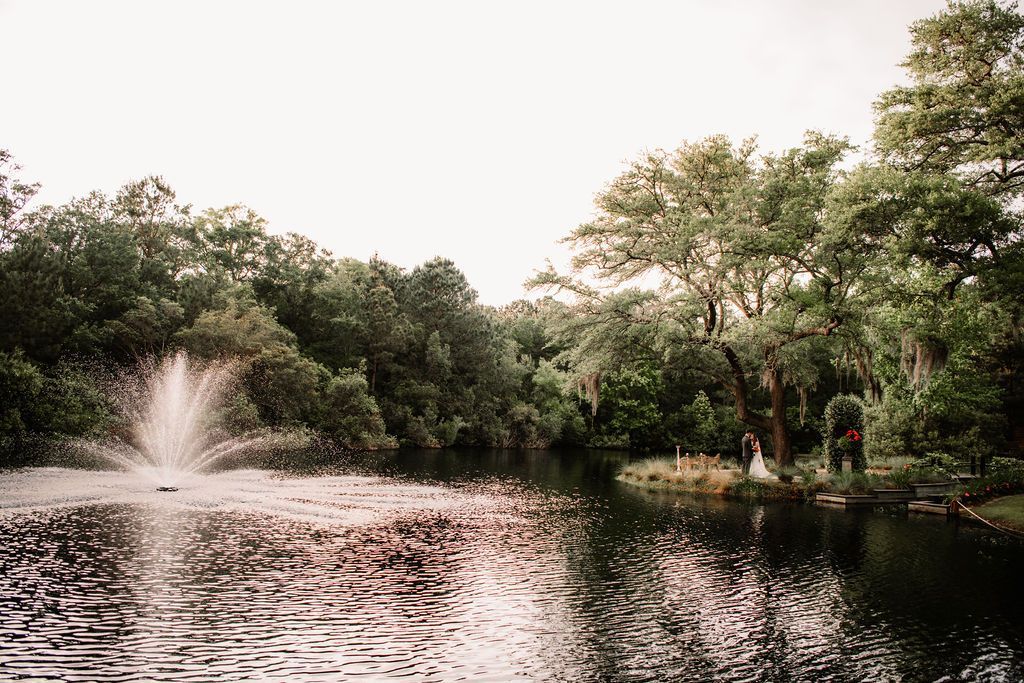 Serene pond with a fountain, surrounded by lush green trees and a few people on the far bank.