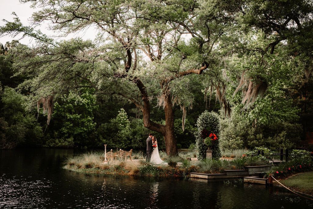 Bride and groom standing by a pond in a lush garden under tall trees