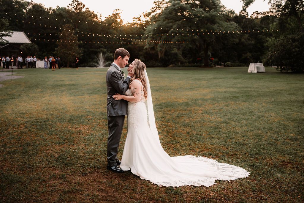 Couple in wedding attire embracing on a grassy lawn at sunset, bride's white train flowing behind.