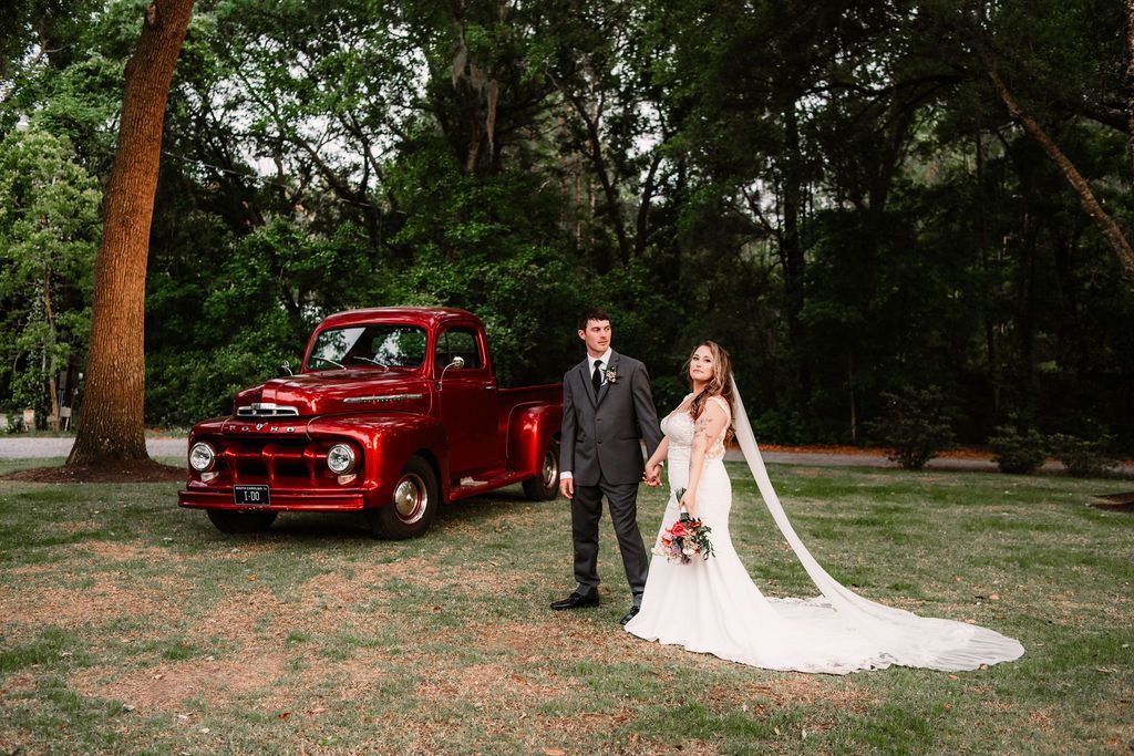Bride and groom standing beside a vintage red truck in a wooded outdoor setting