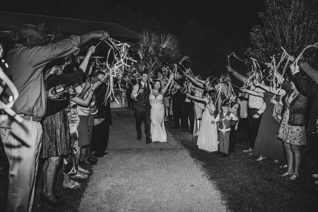 Crowd cheers under an arch of raised arms as a couple walks down a lit outdoor path at night
