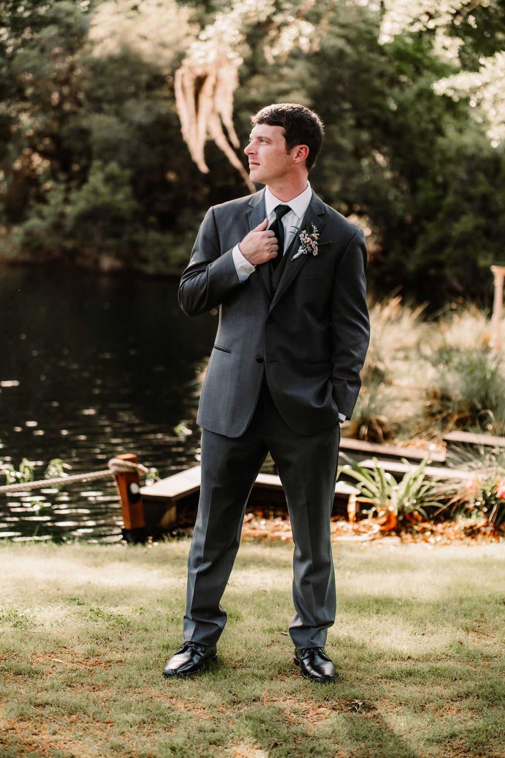 Man in a dark suit adjusting his tie outdoors in a garden with trees and sunlight