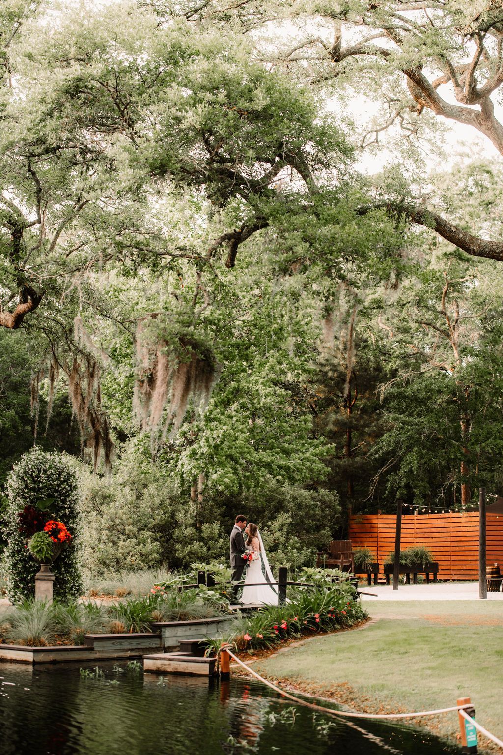 Tree-filled park with a small bridge, pond, and a person walking on a path near orange fencing