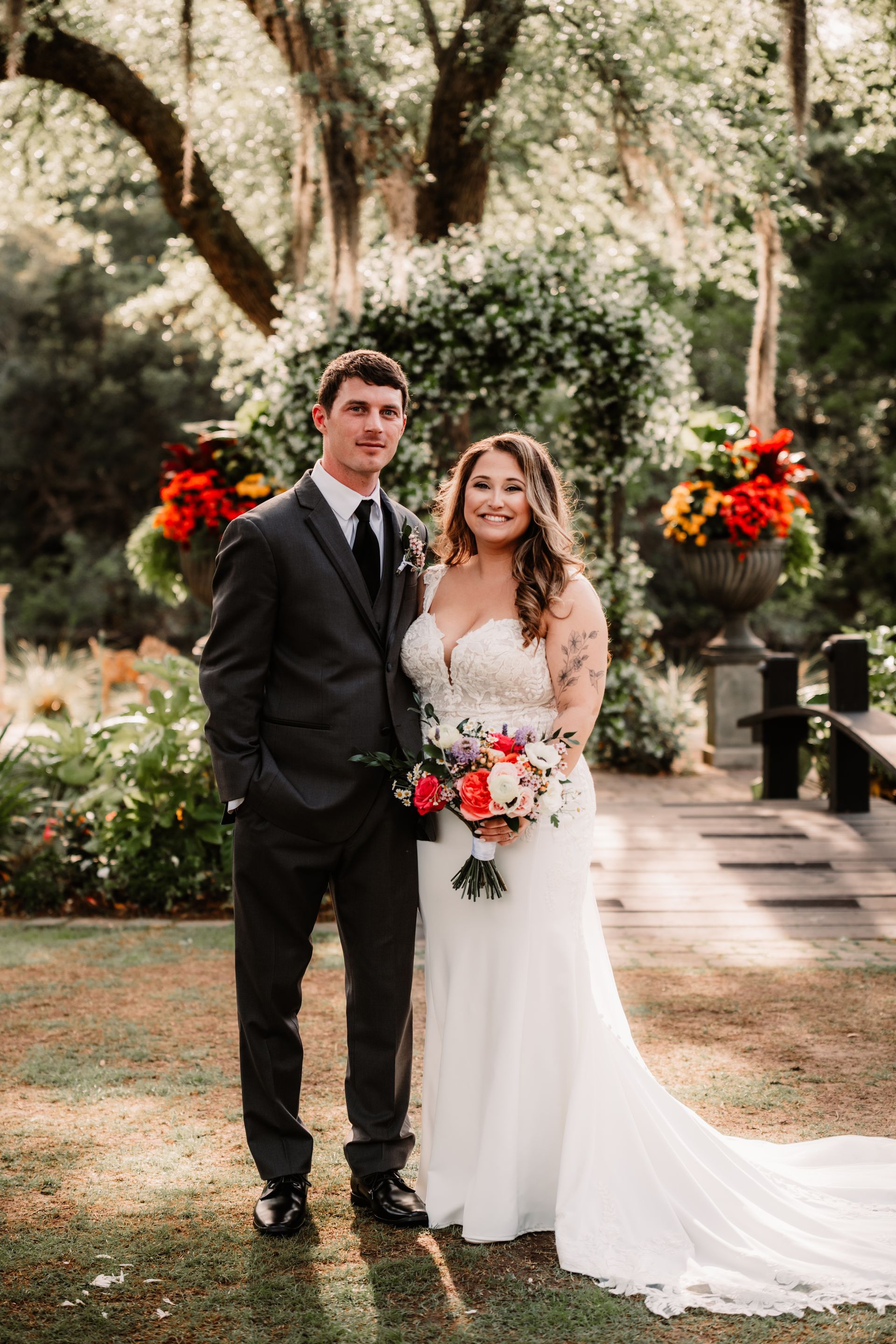 Couple in wedding attire posing outdoors in a garden, holding a red-and-white bouquet.