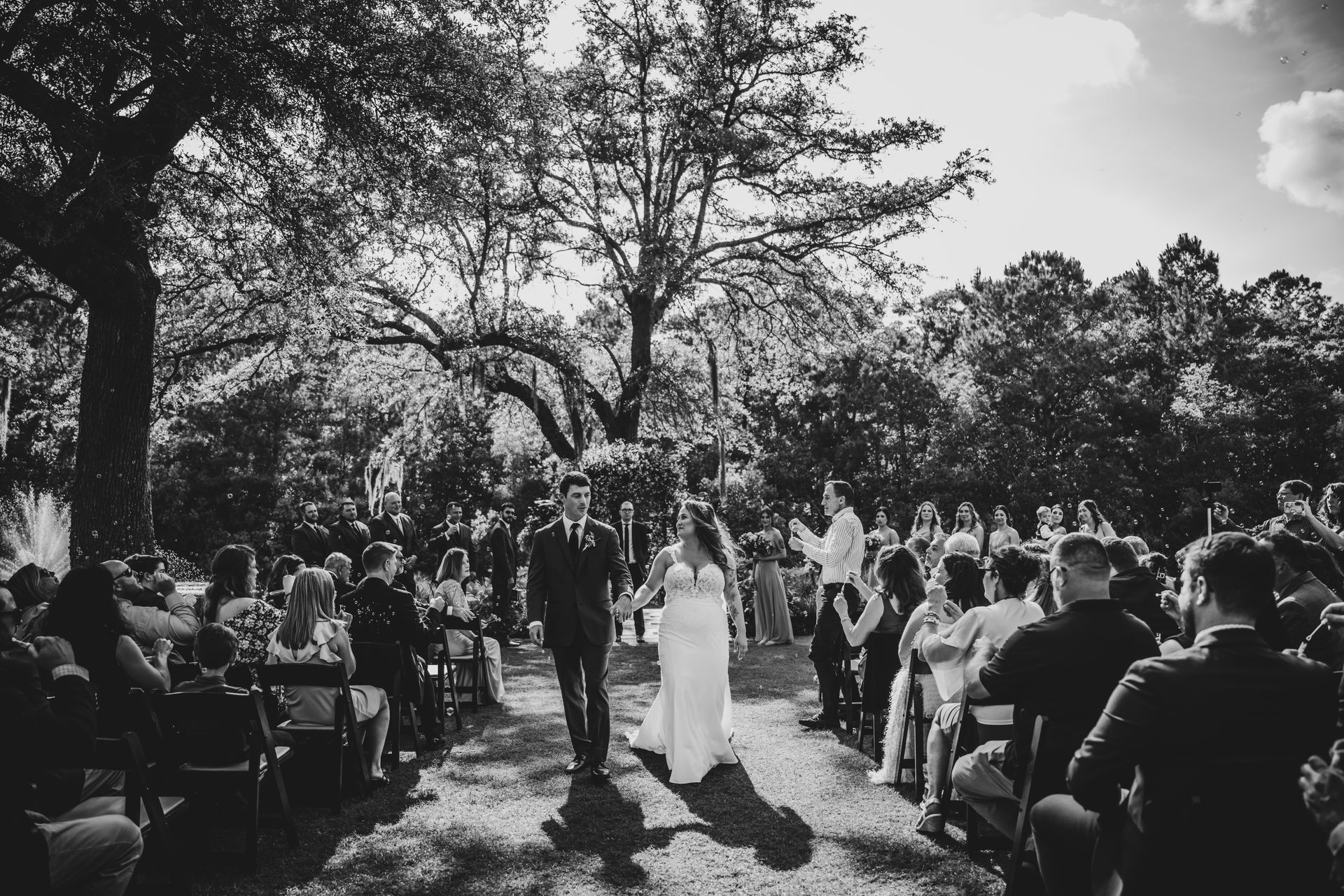 Outdoor wedding ceremony under trees, with bride and groom walking between seated guests.