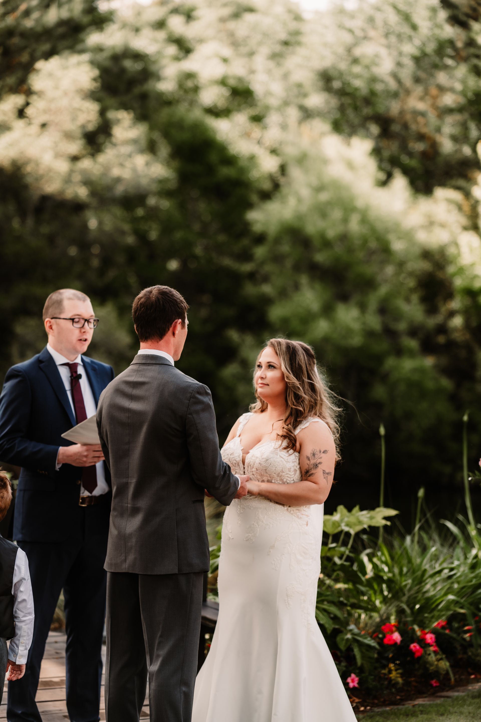 Wedding couple exchanging vows outdoors with officiant, greenery and flowers in background