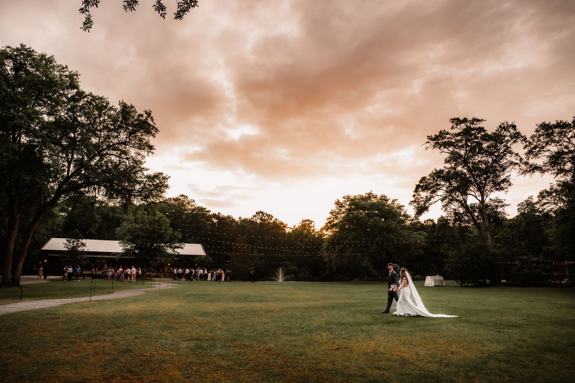 Bride and groom standing on a grassy field at sunset, framed by trees and a warm orange sky.