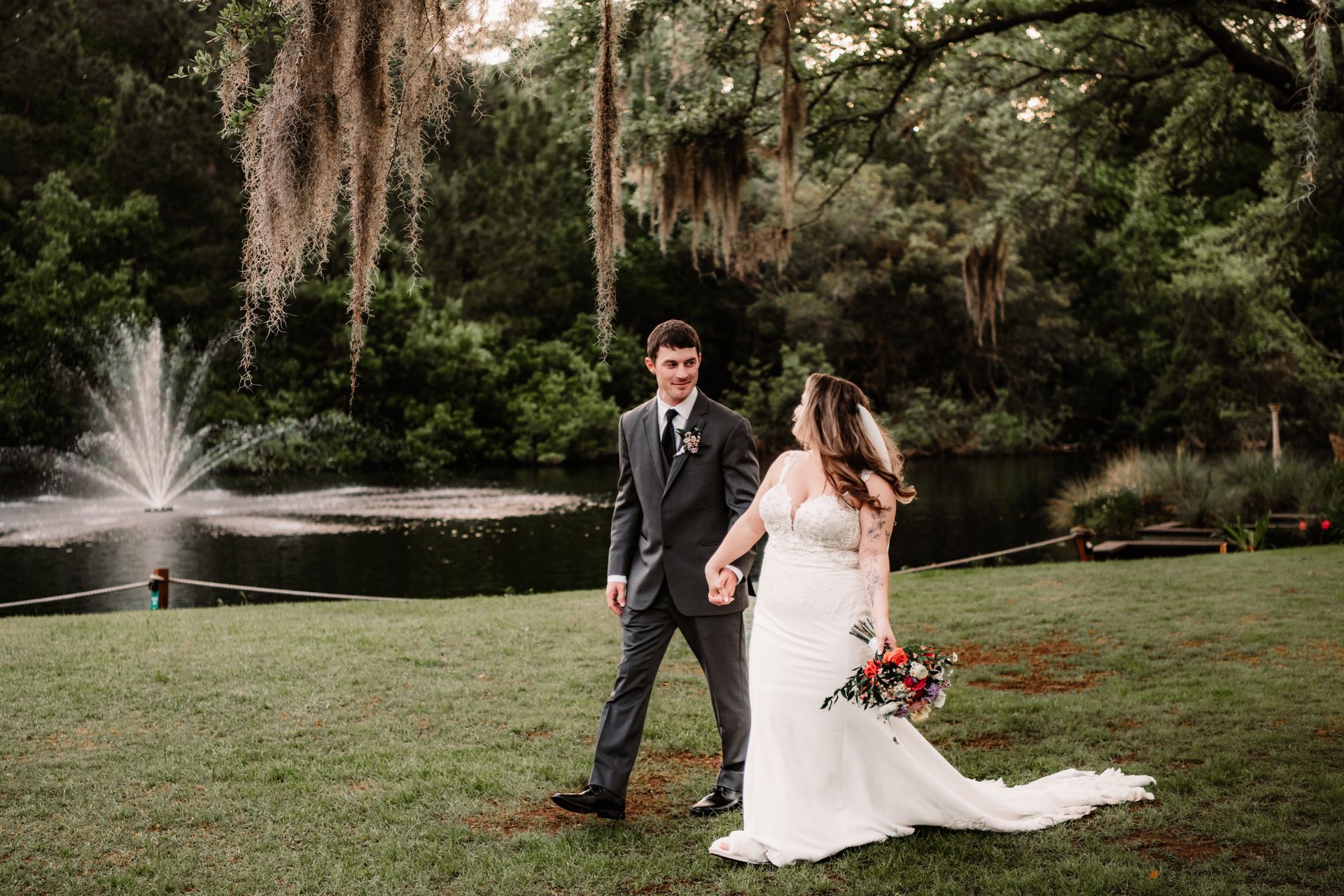 Bride and groom walking on grass beside a lake fountain in a wooded park
