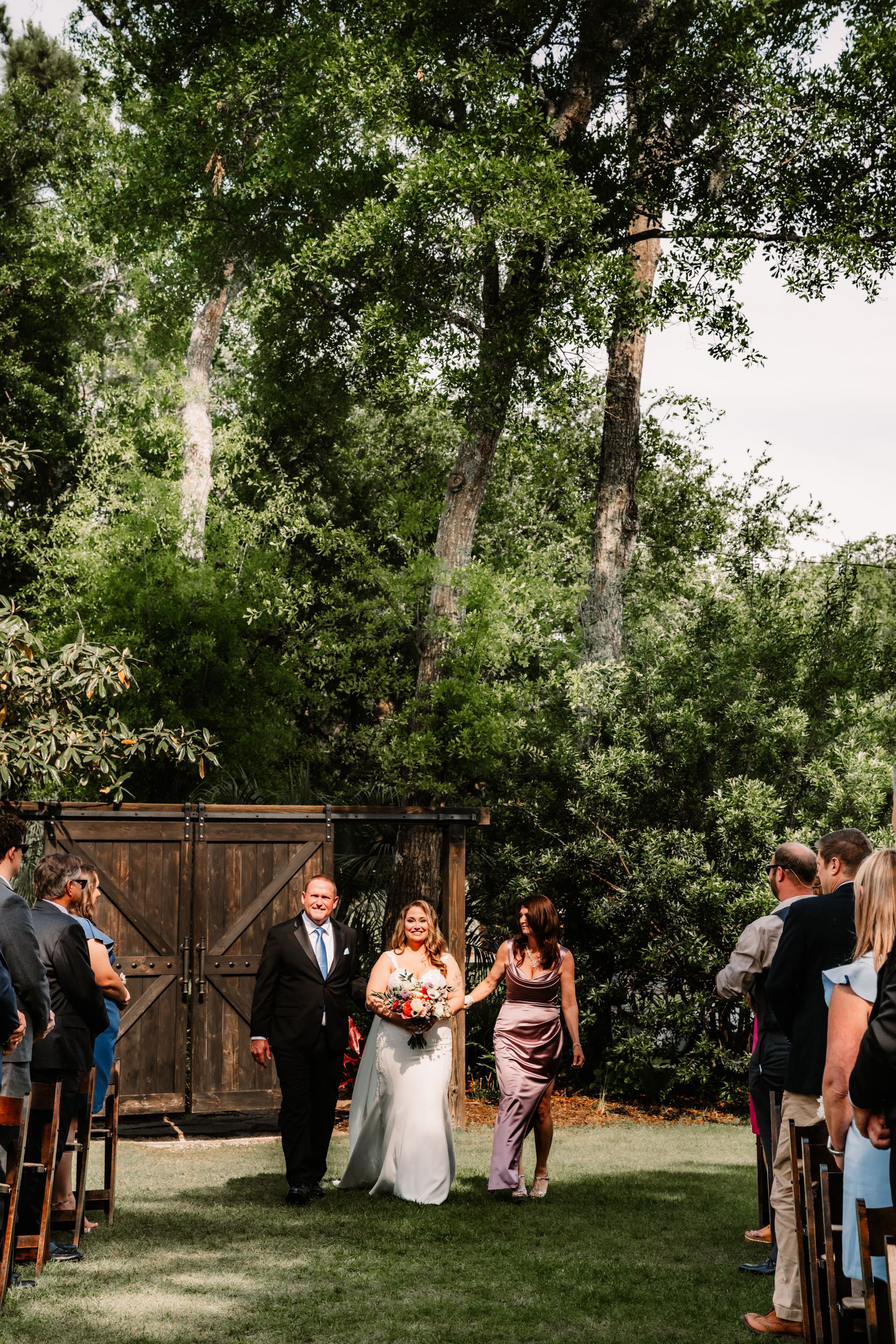 Bride and groom walk down an outdoor aisle under trees, guests seated on both sides.