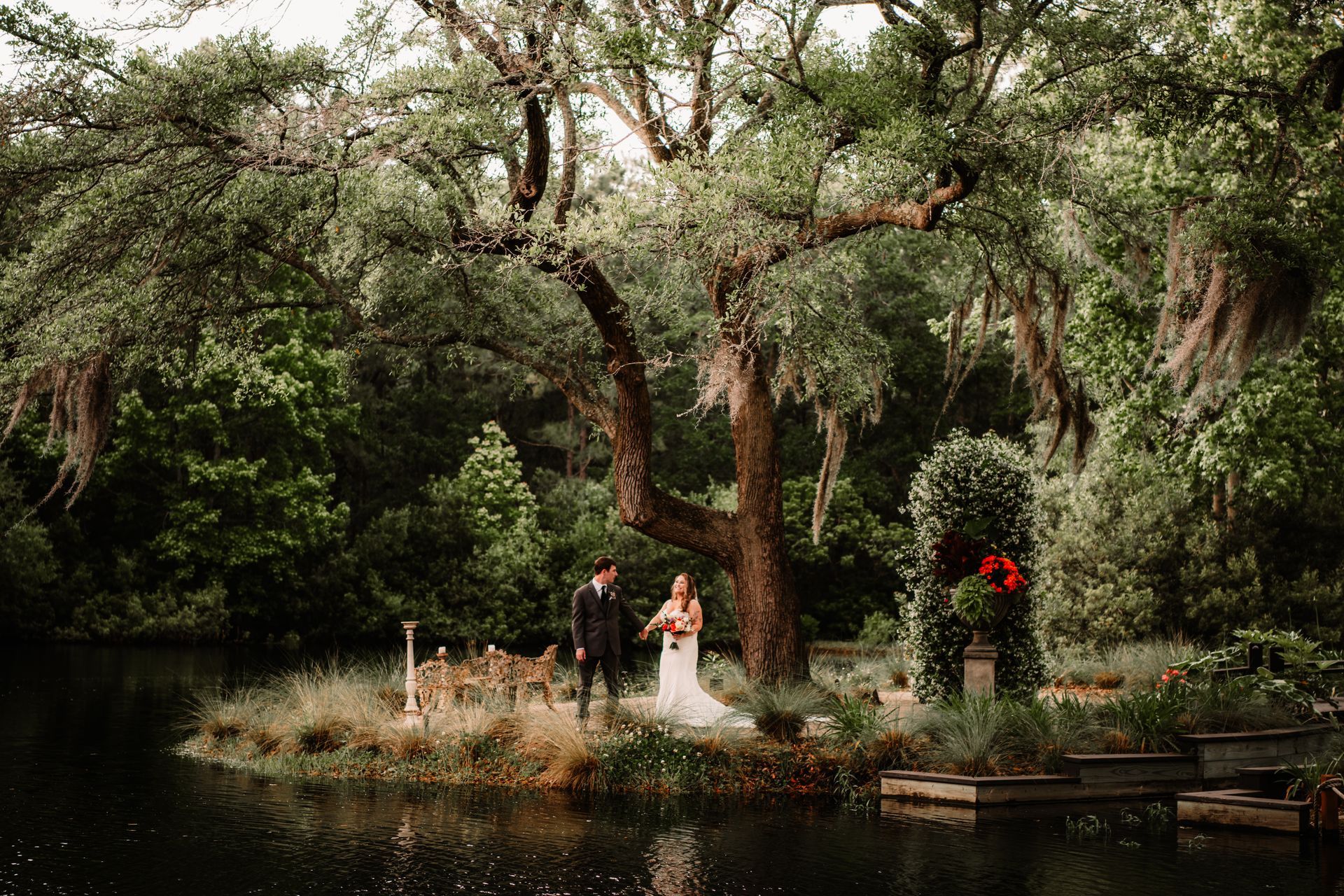 Wedding couple by a pond in a lush forest garden, surrounded by trees and greenery.