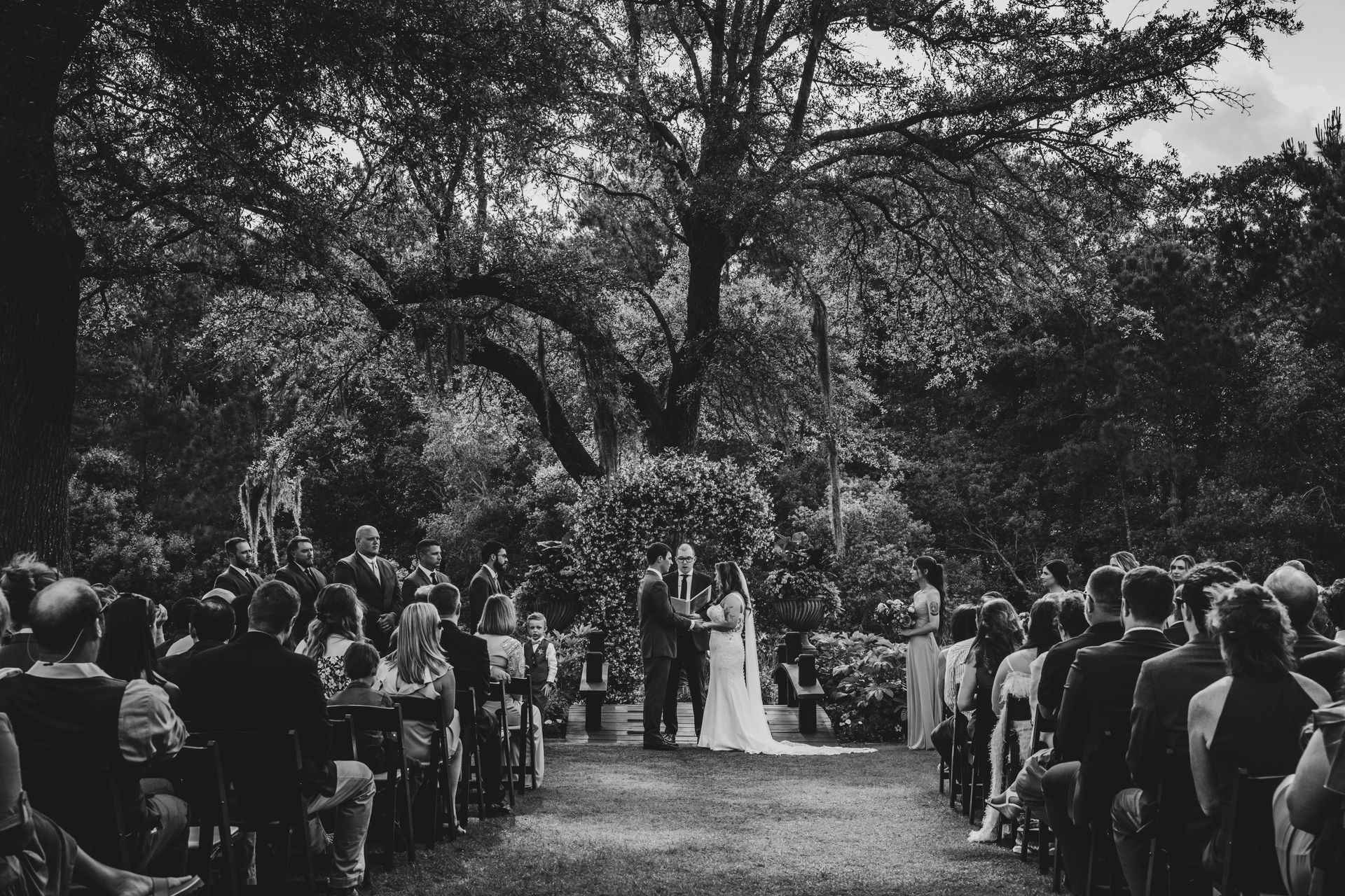 Outdoor wedding ceremony under large trees, with guests seated along a garden aisle.