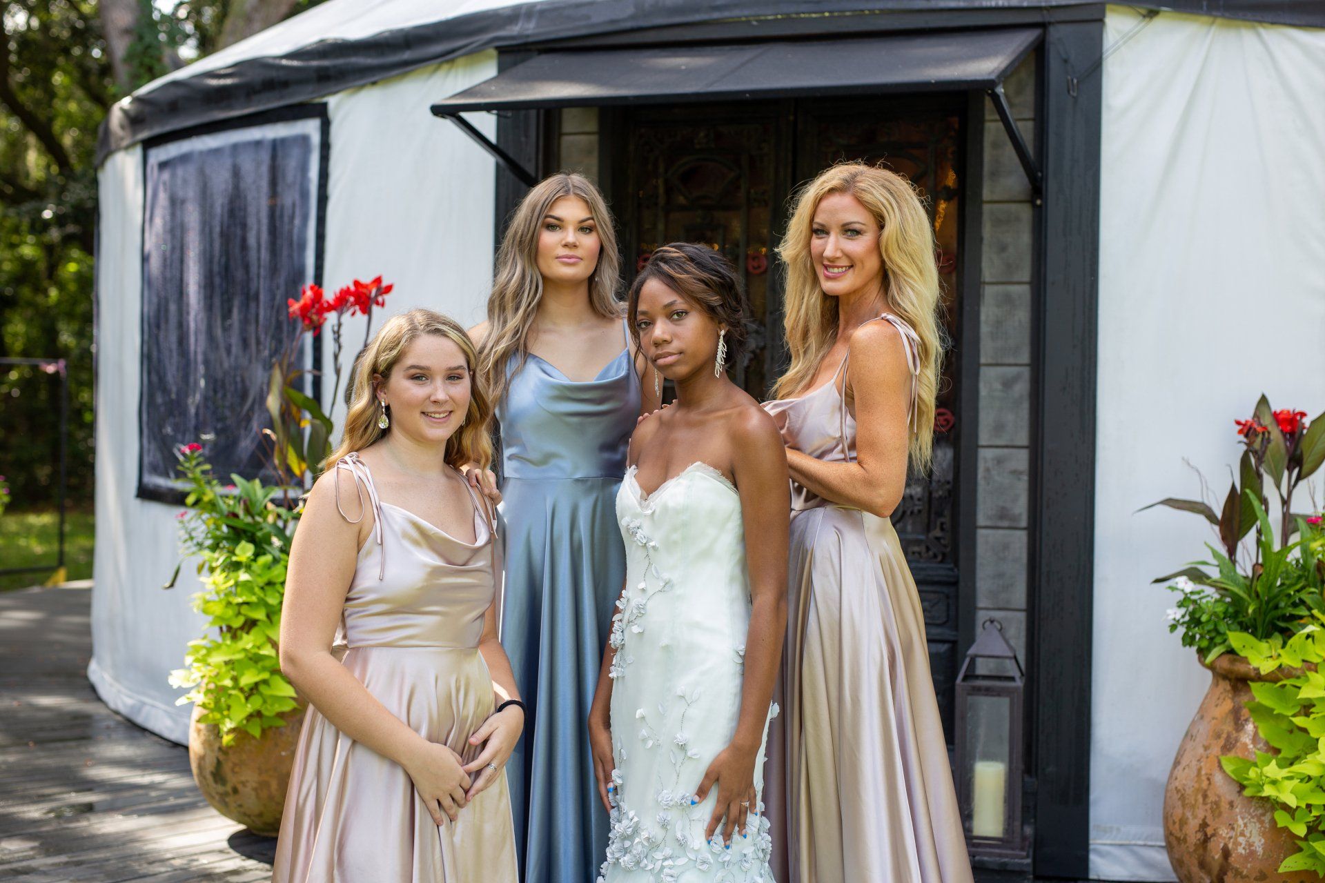 Four women in formal dresses posing outside a dark tent with flowers and greenery.