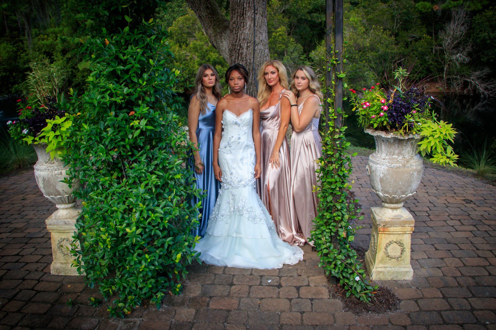 Four women in formal gowns stand on a brick path framed by lush greenery and stone urns.