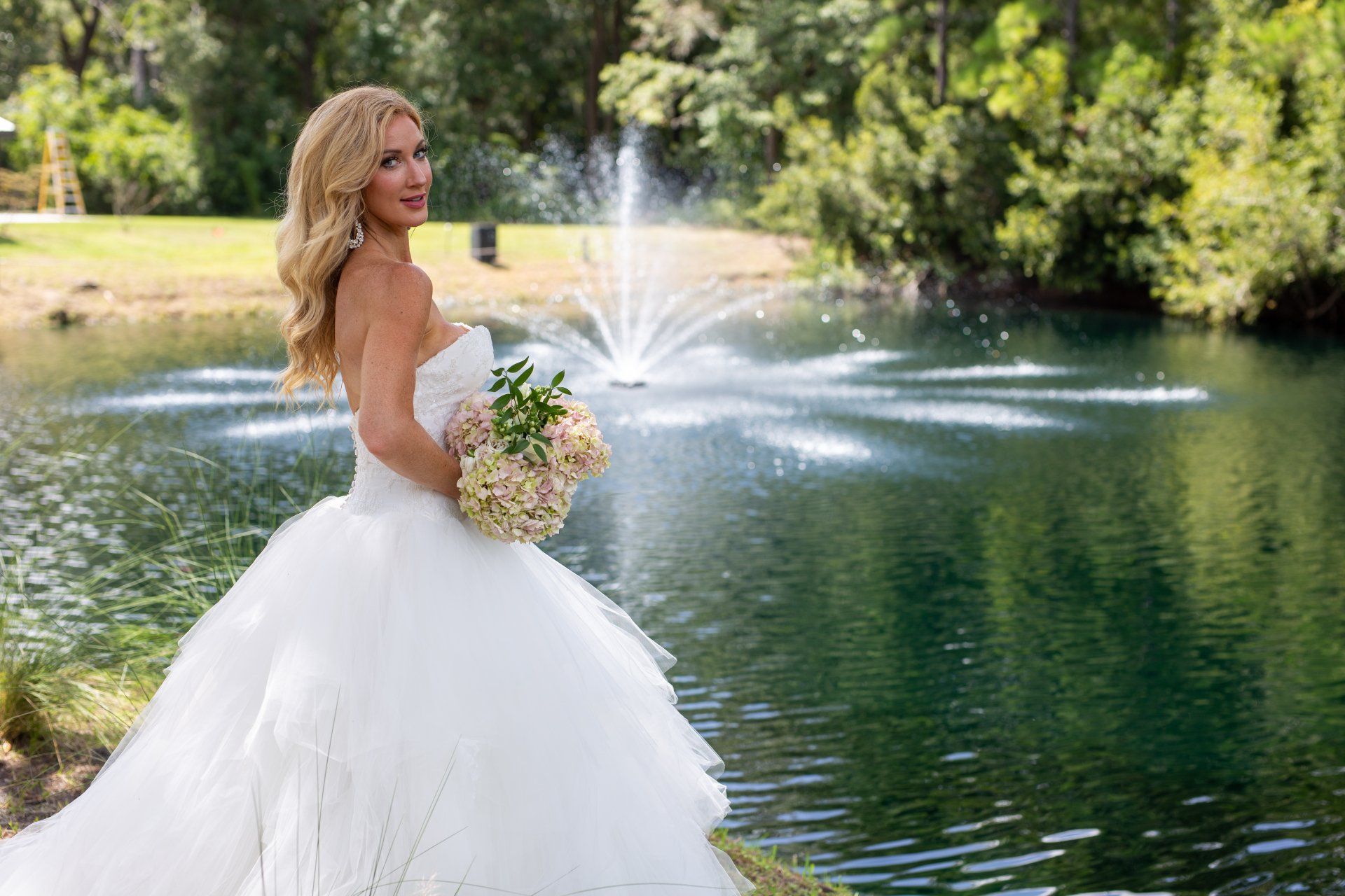 Bride in white gown holding bouquet by a lake fountain, with trees in the background