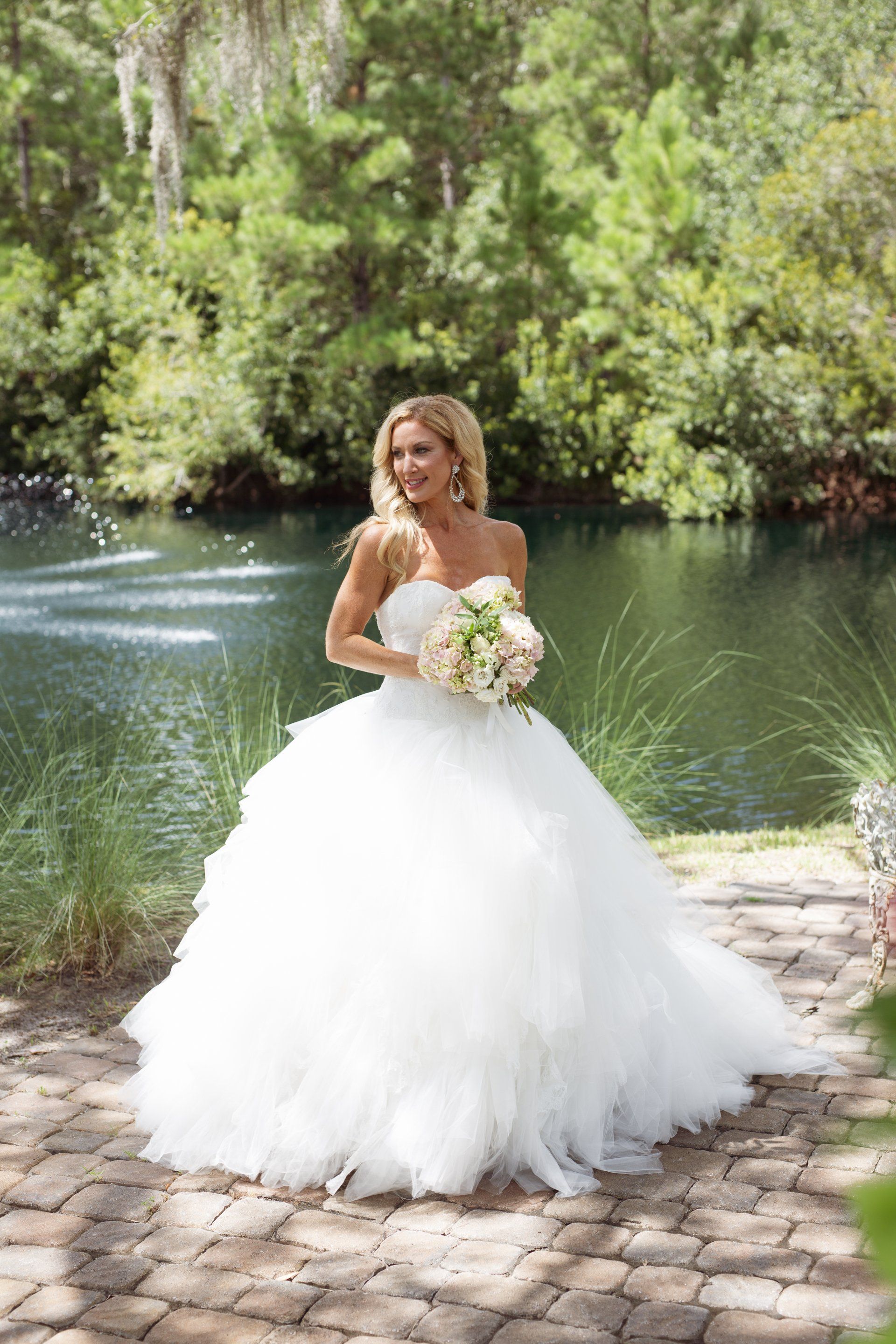 Bride in a white gown holding a bouquet beside a pond with a fountain and trees