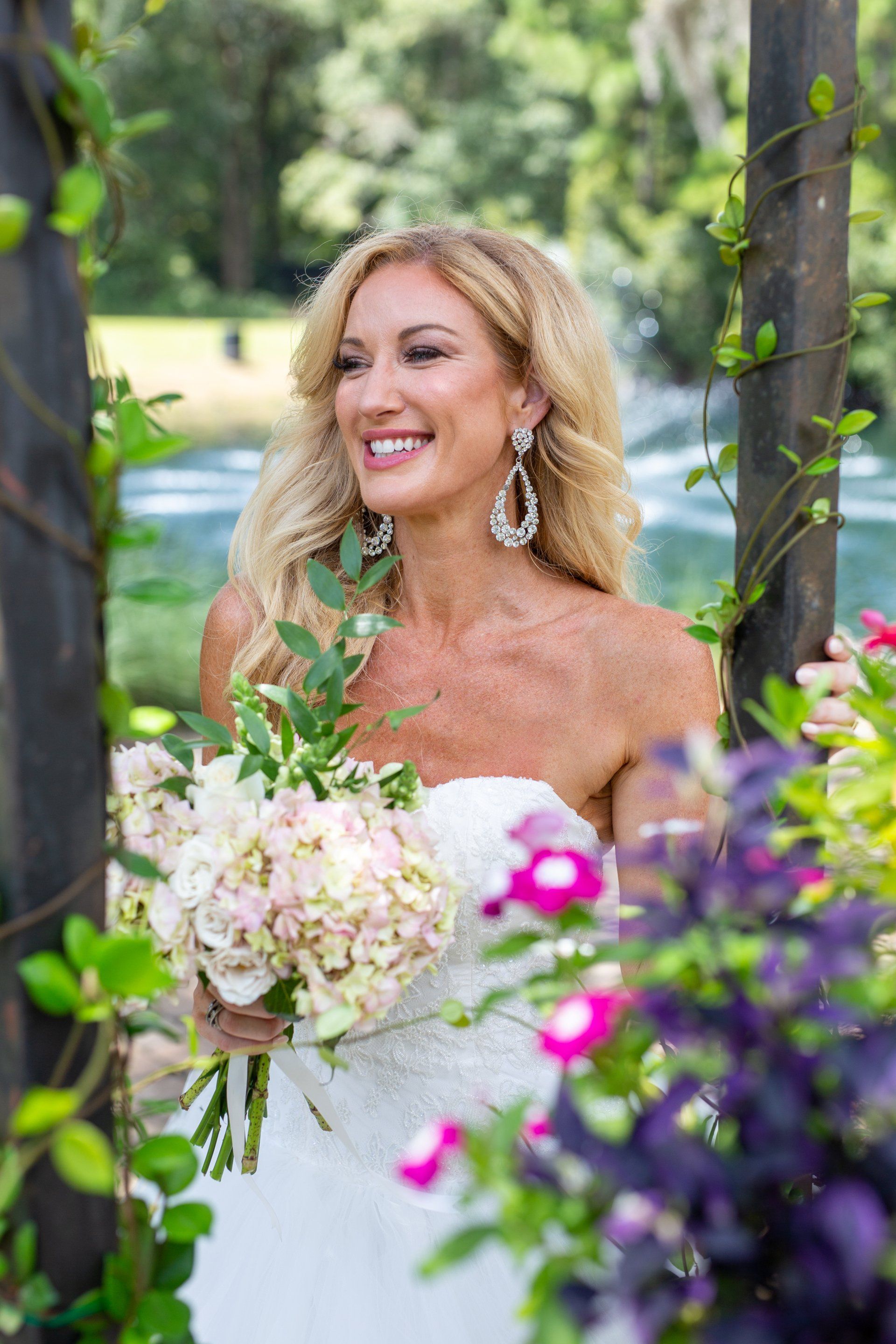 Woman in a white dress holding a bouquet in a garden, smiling beside a tree.