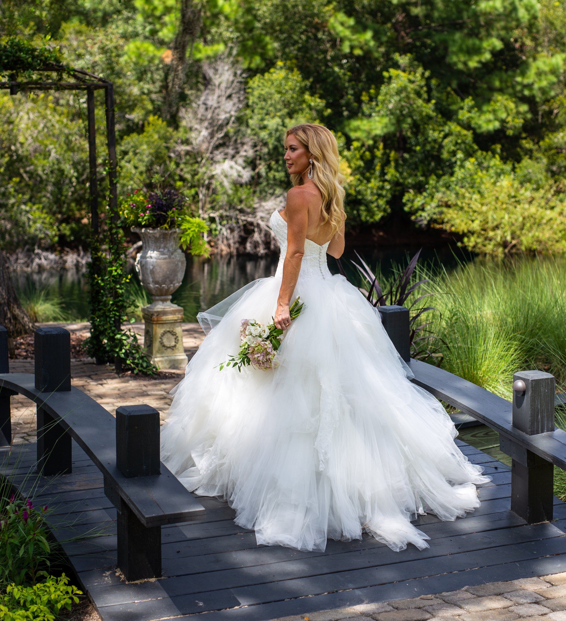 Bride in a white gown standing on a garden bridge with greenery and flowers surrounding her