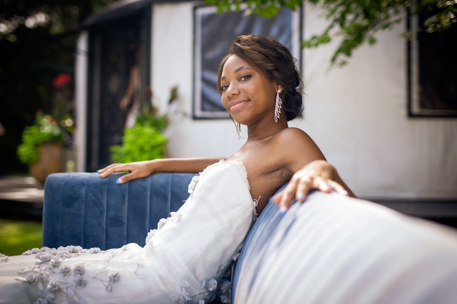 Bride in a white dress reclining on a blue couch, smiling outdoors with greenery in the background