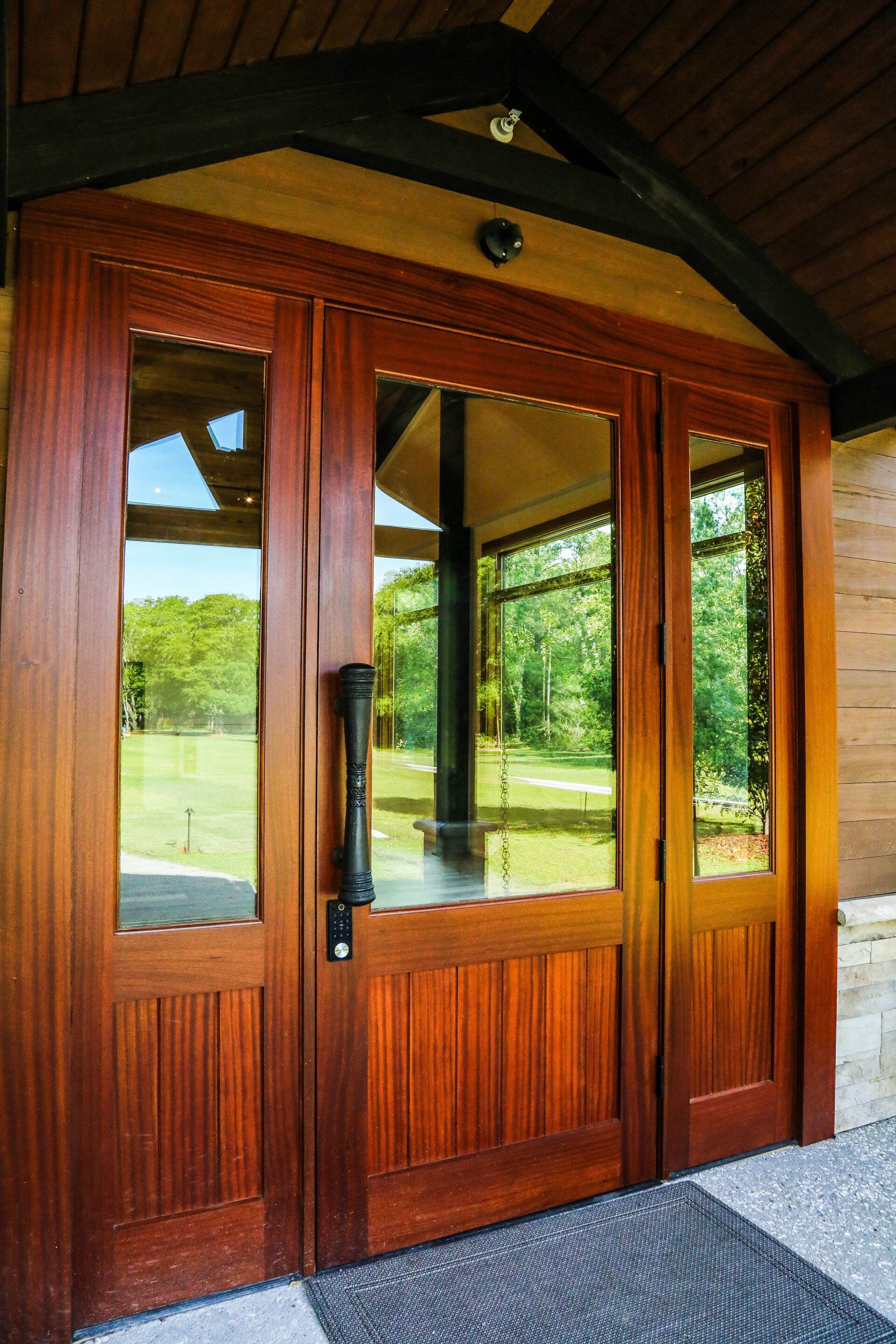 Wooden double glass doors under a covered entrance, with reflections of greenery outside.