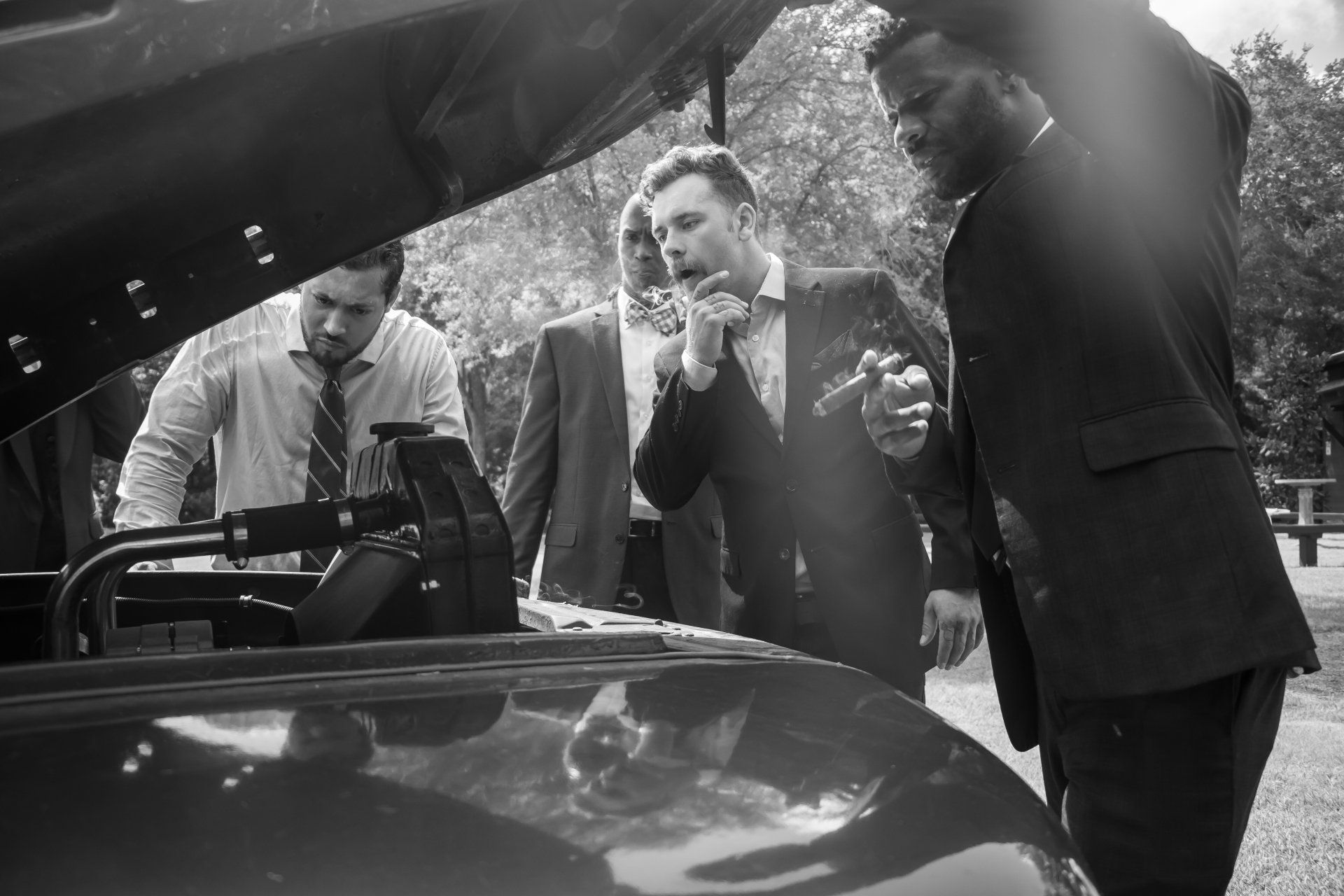 Three men examine a car engine outdoors, with one holding a cigarette in black-and-white.