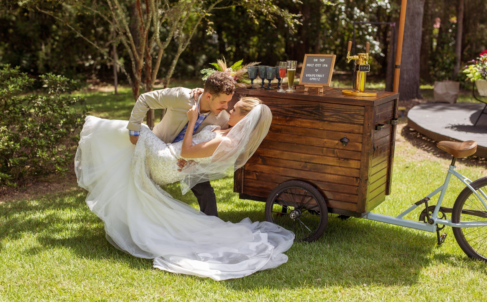 Bride in a white dress leaning against a wooden cart in a sunny garden setting