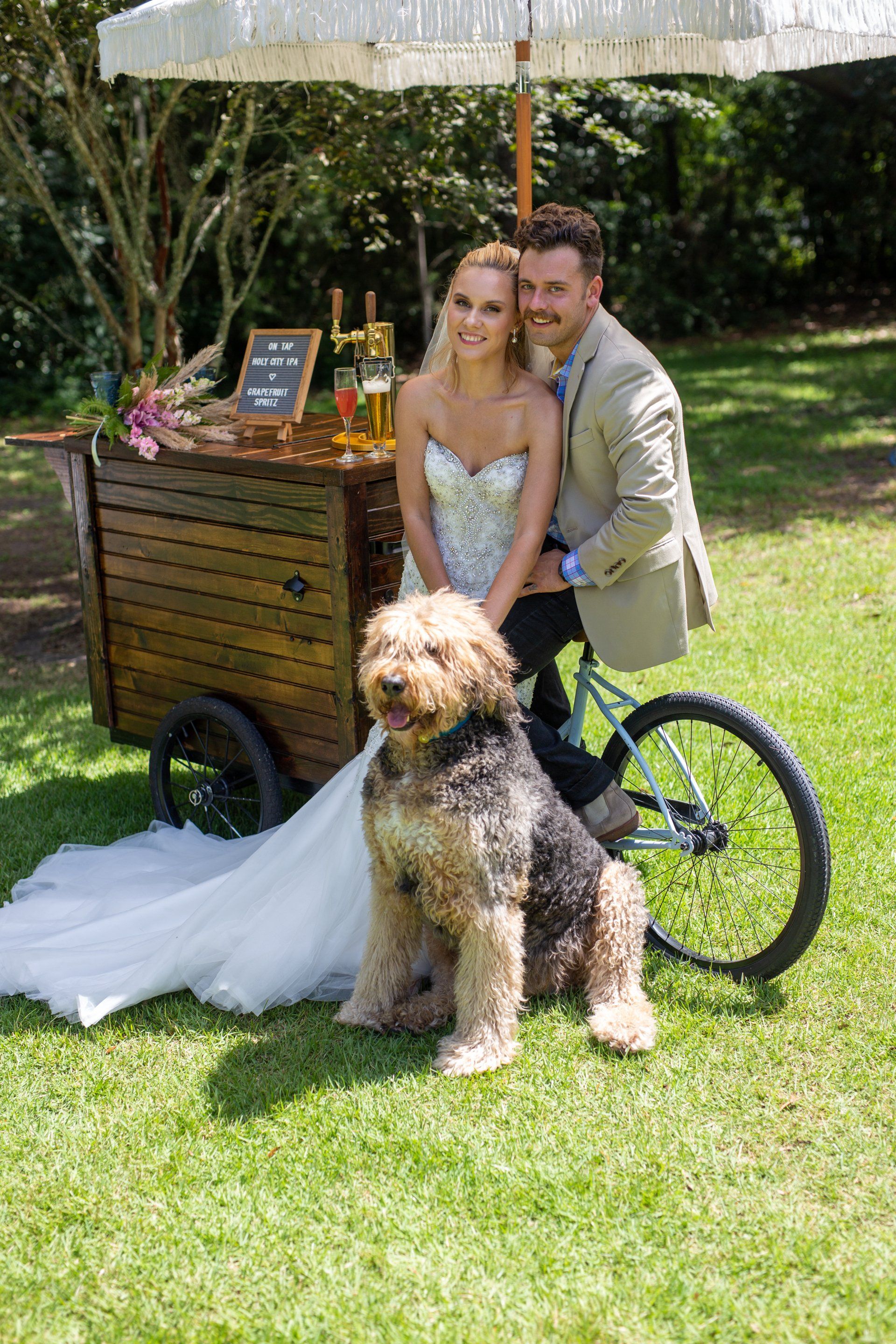 Couple posing with a fluffy dog beside a wooden cart under a white canopy on a sunny lawn