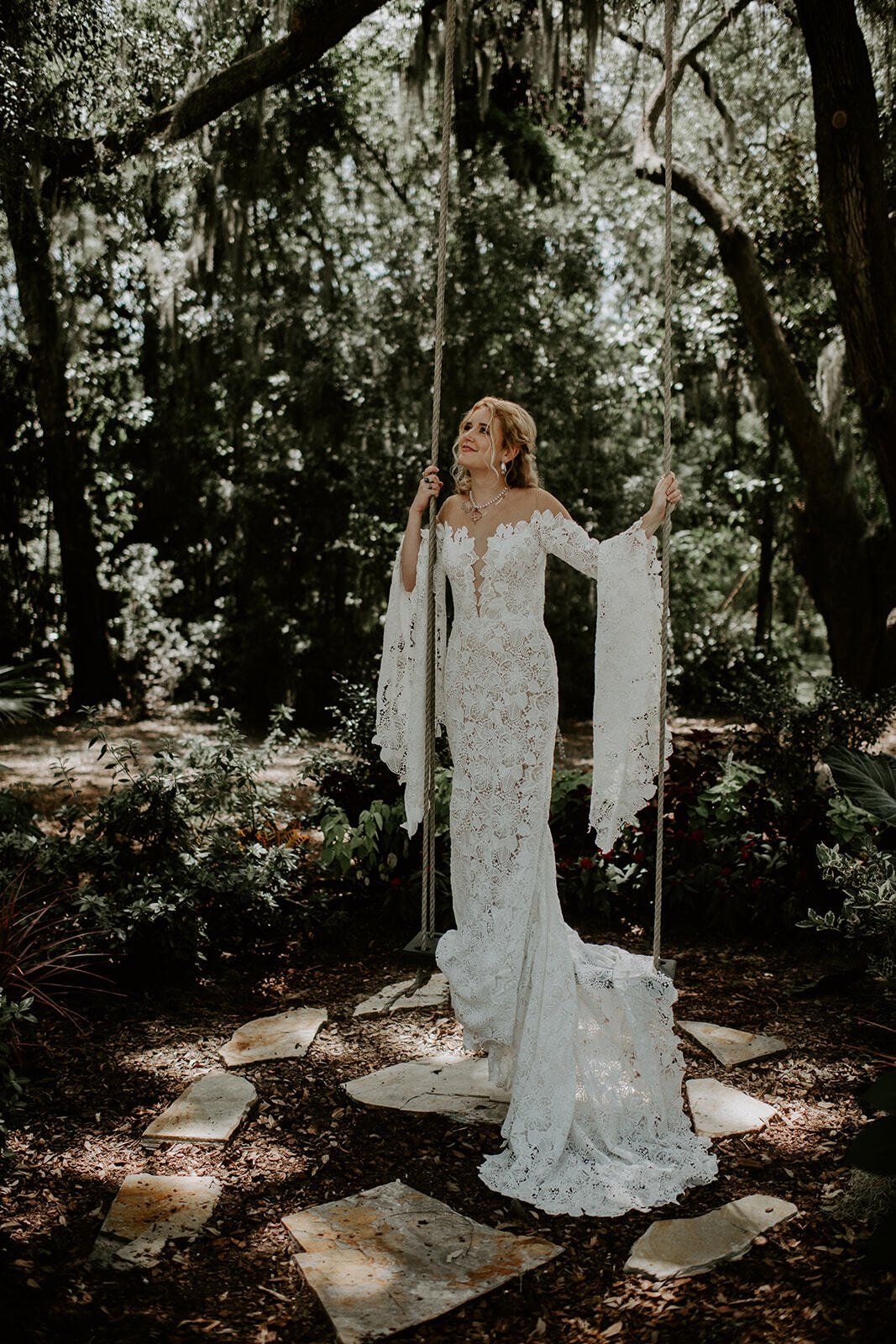 White-gowned figure standing in a sunlit wooded path with flowing veil and raised arms