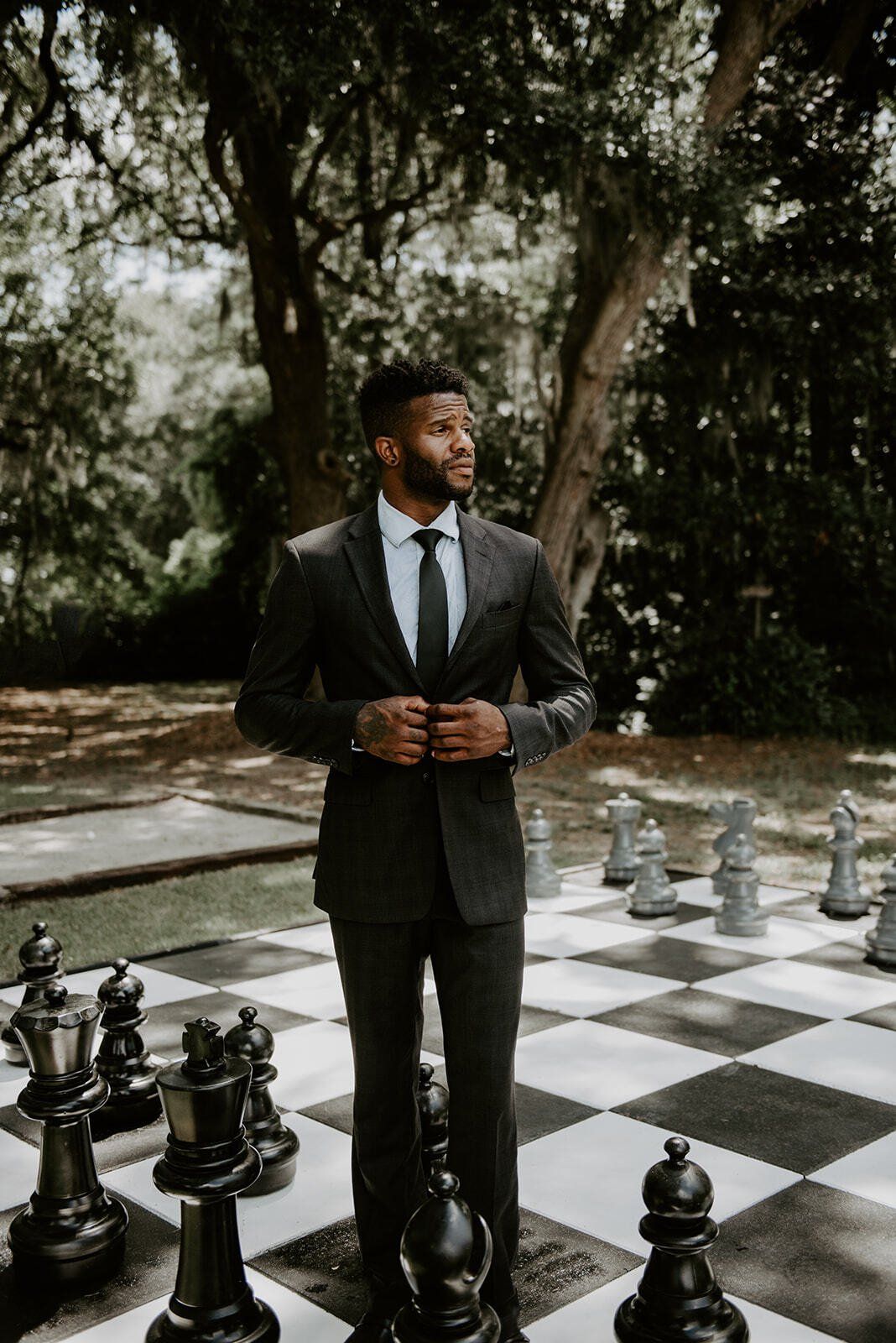 Man in a dark suit standing on a giant outdoor chessboard in a garden