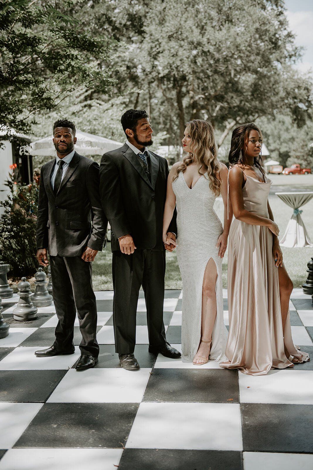 Four people in formal attire standing on a black-and-white checkered floor outdoors under trees