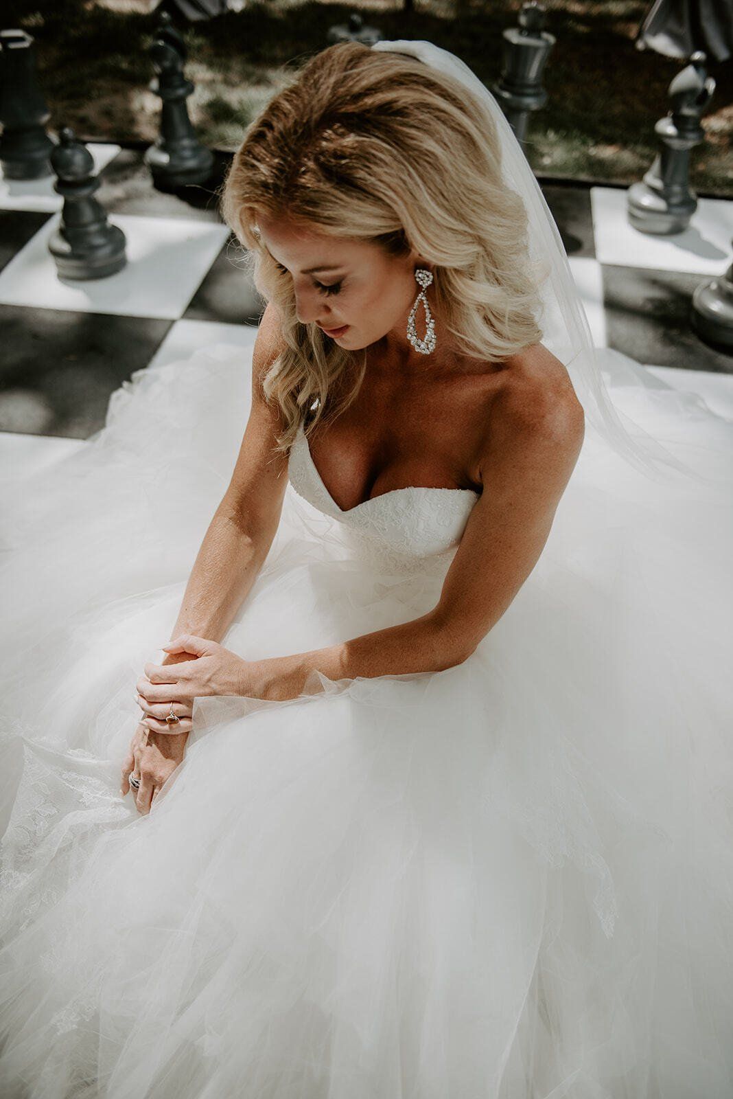 Bride in a white strapless gown sitting on a checkered floor, adjusting her hands and looking down.