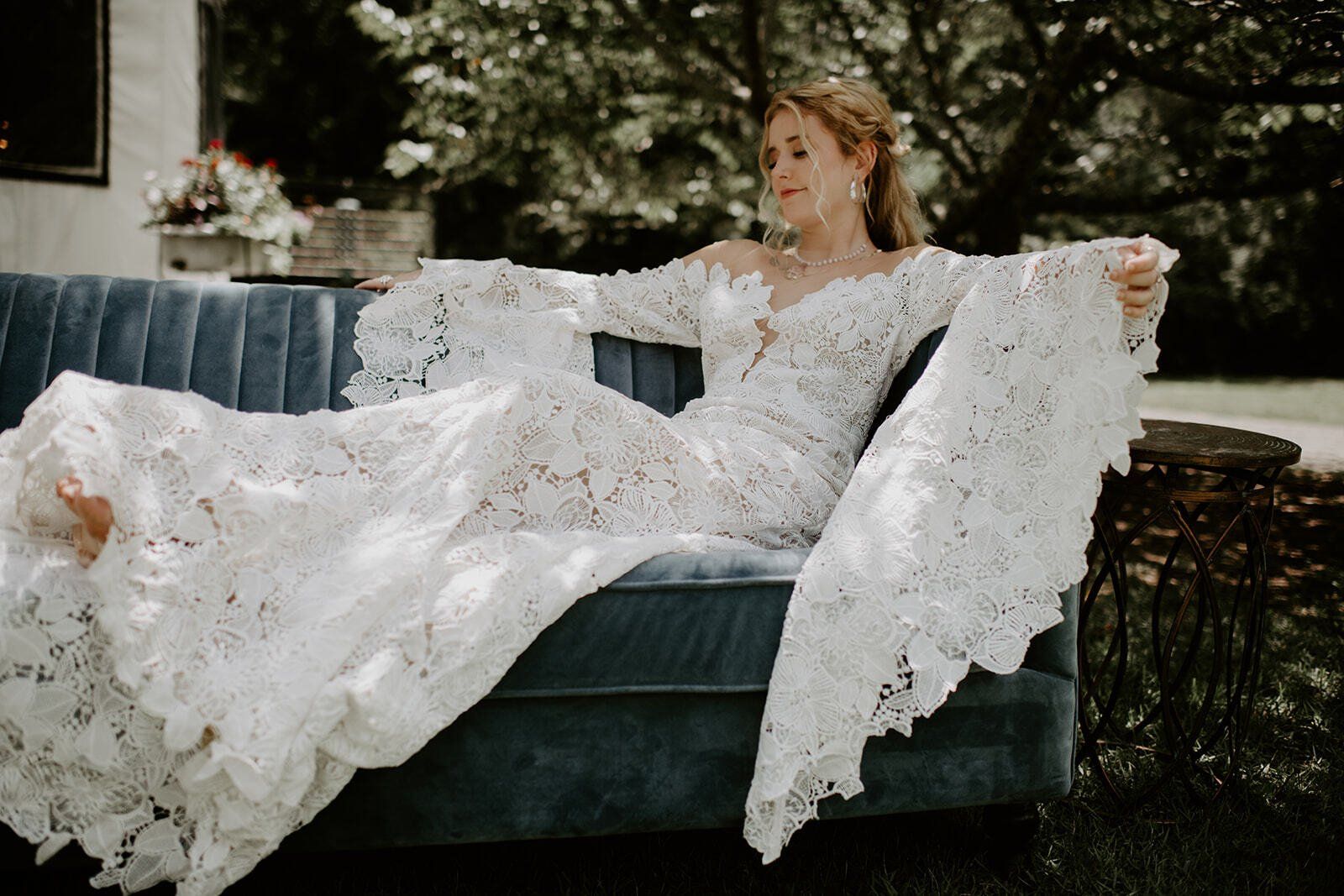 Woman in a white lace dress reclining on a teal couch outdoors, with a wicker chair nearby.