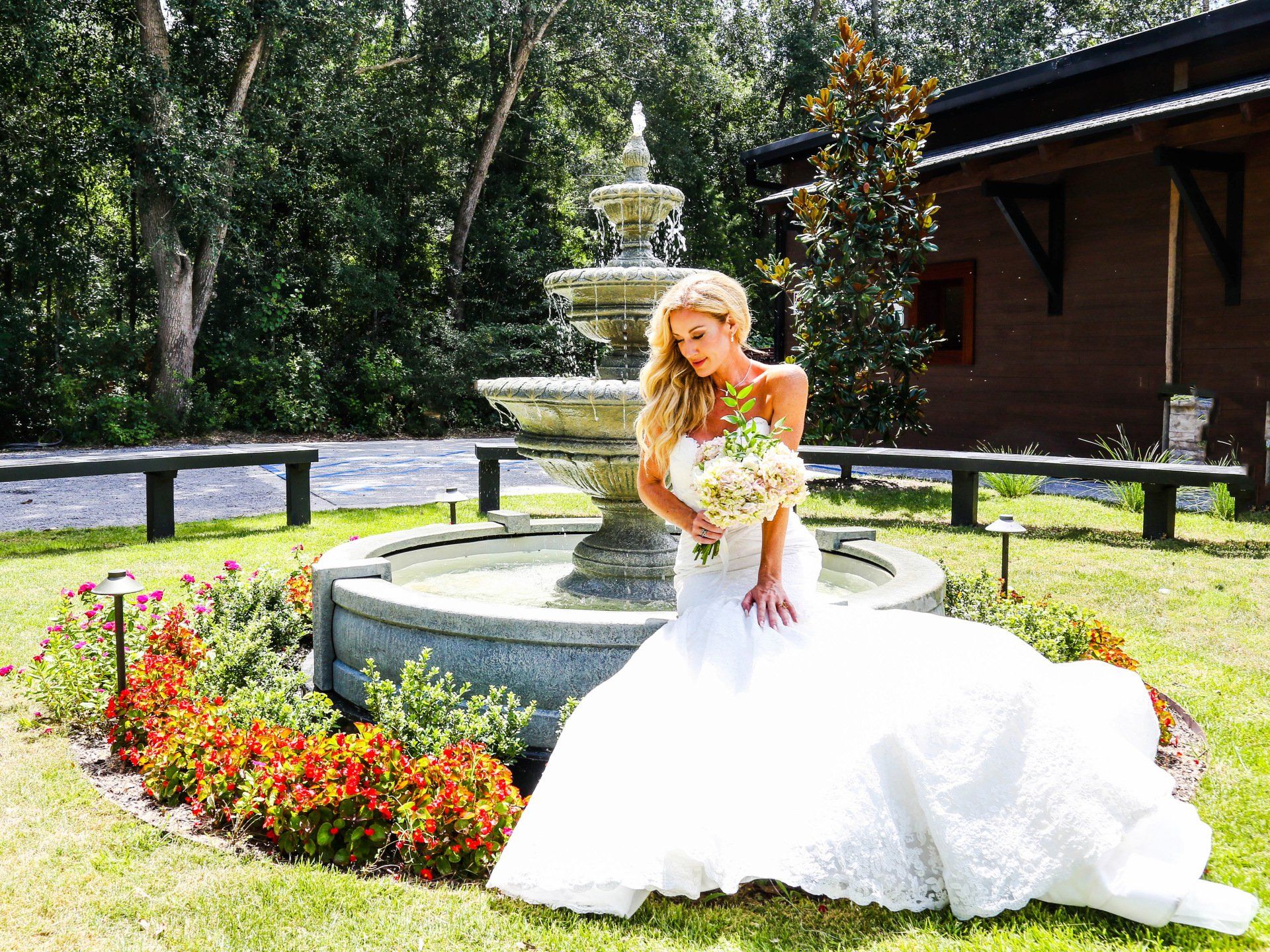 Bride in a white gown holding a bouquet beside a stone fountain and flower bed in a garden