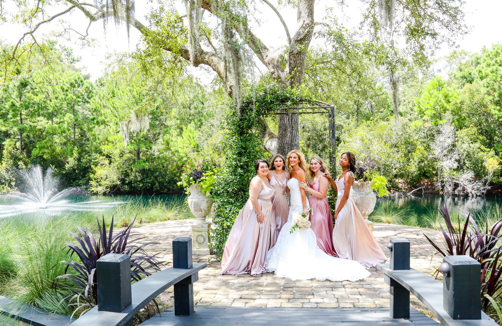 Bridal party in pink dresses posing on a garden bridge beside a fountain and lush greenery