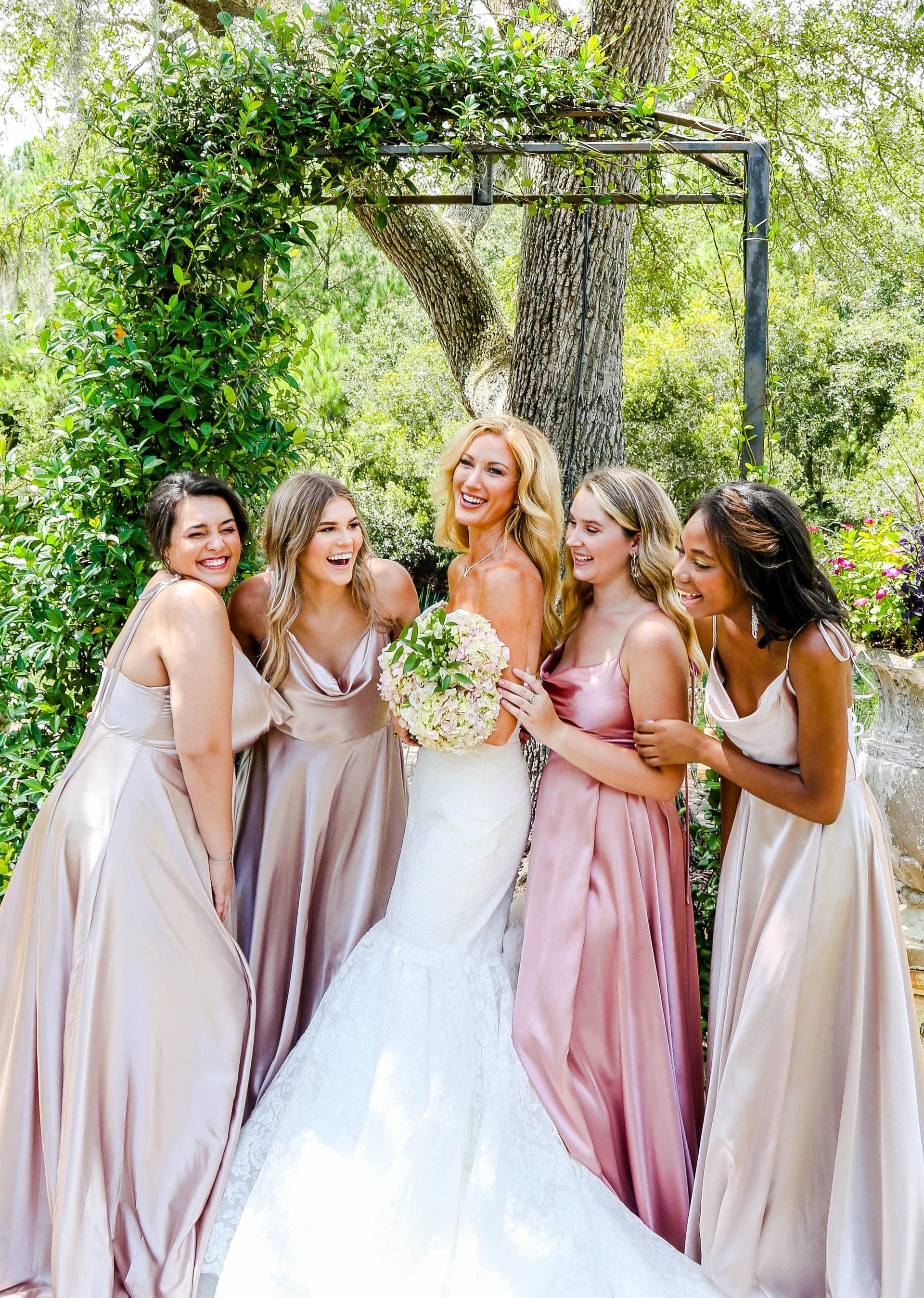 Bride with five bridesmaids in pastel dresses posing outdoors under a floral arch