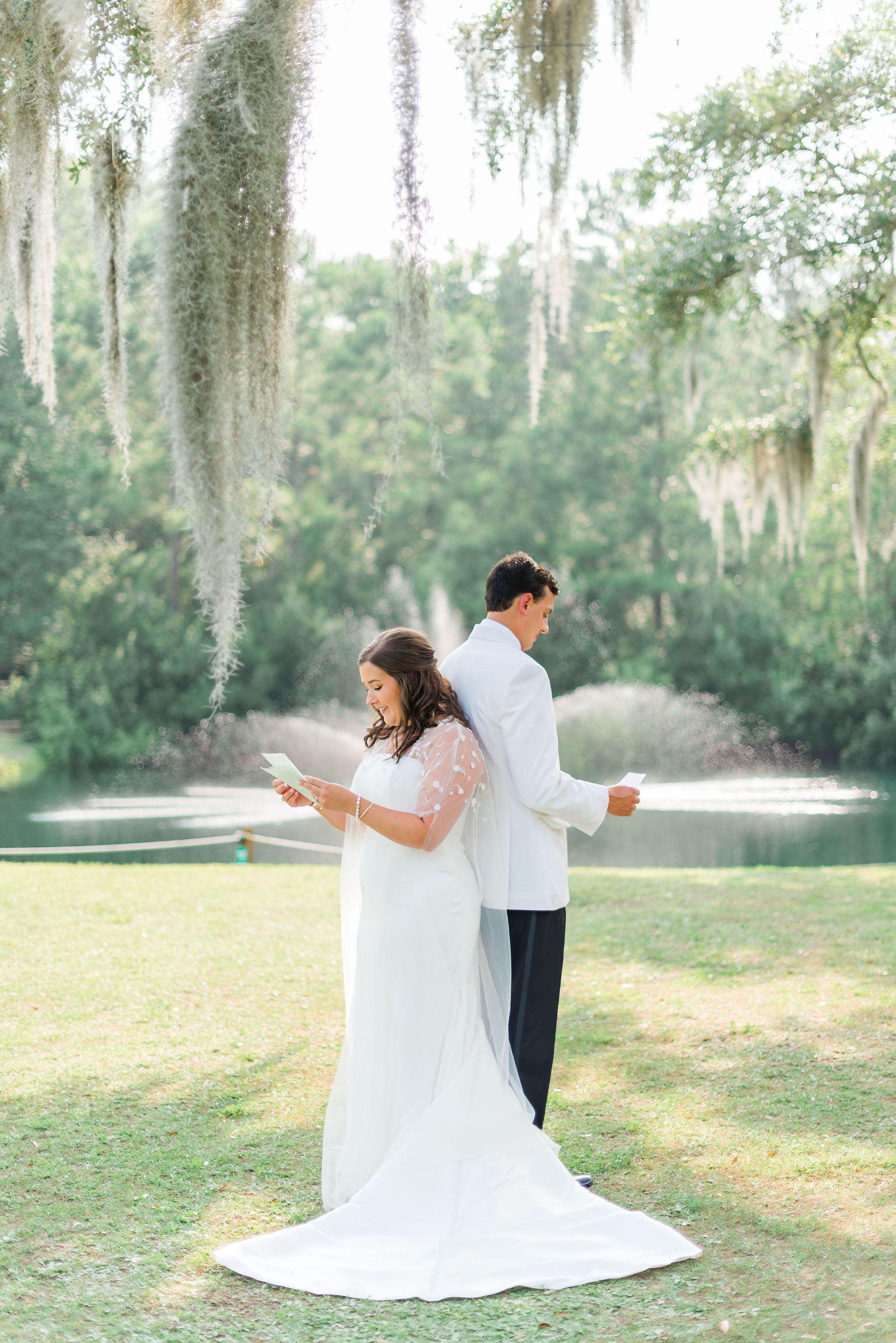 Bride and groom standing back to back in white attire in a sunny garden with fountain and hanging moss