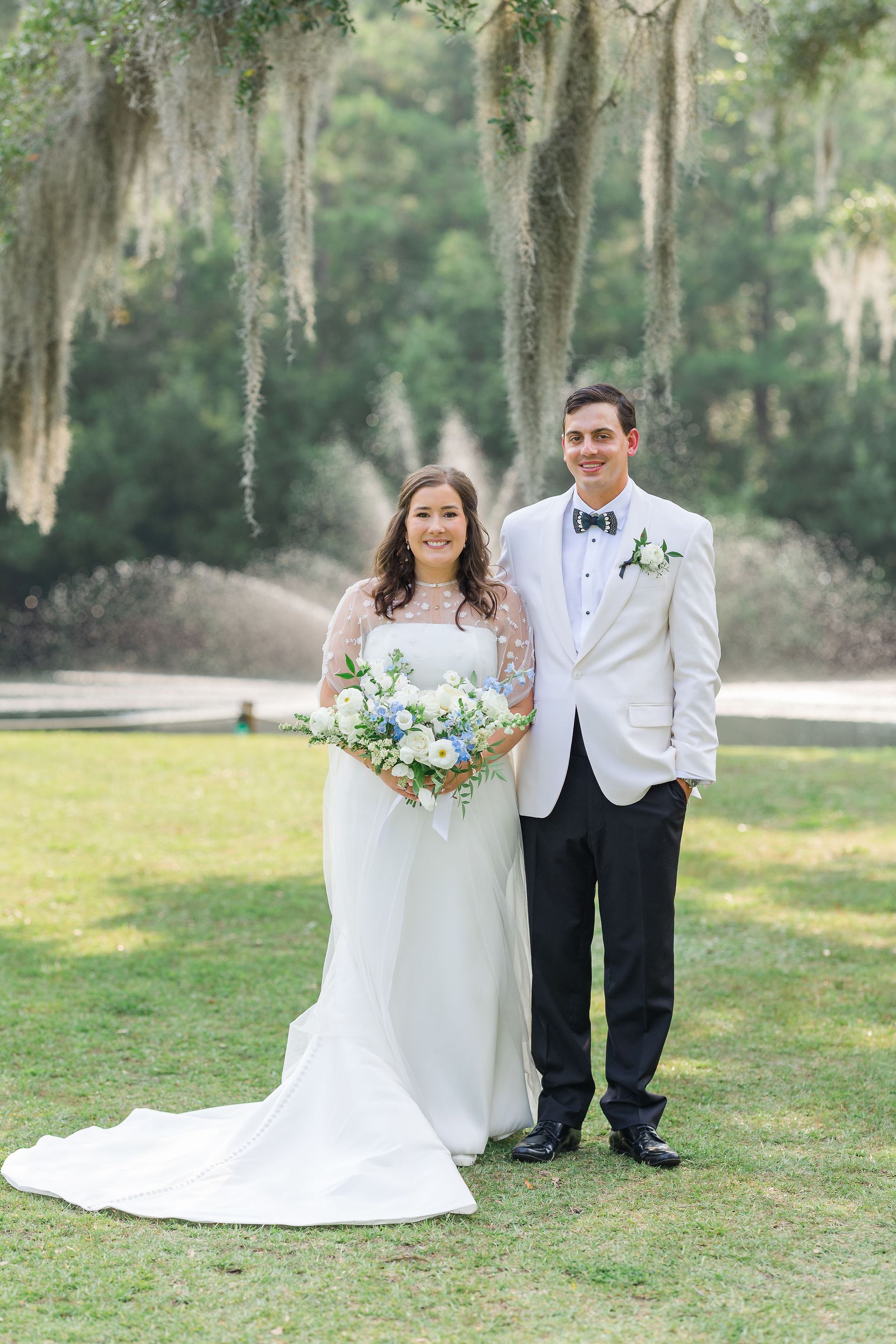 Bride and groom standing together outdoors on grass, smiling and holding a bouquet.