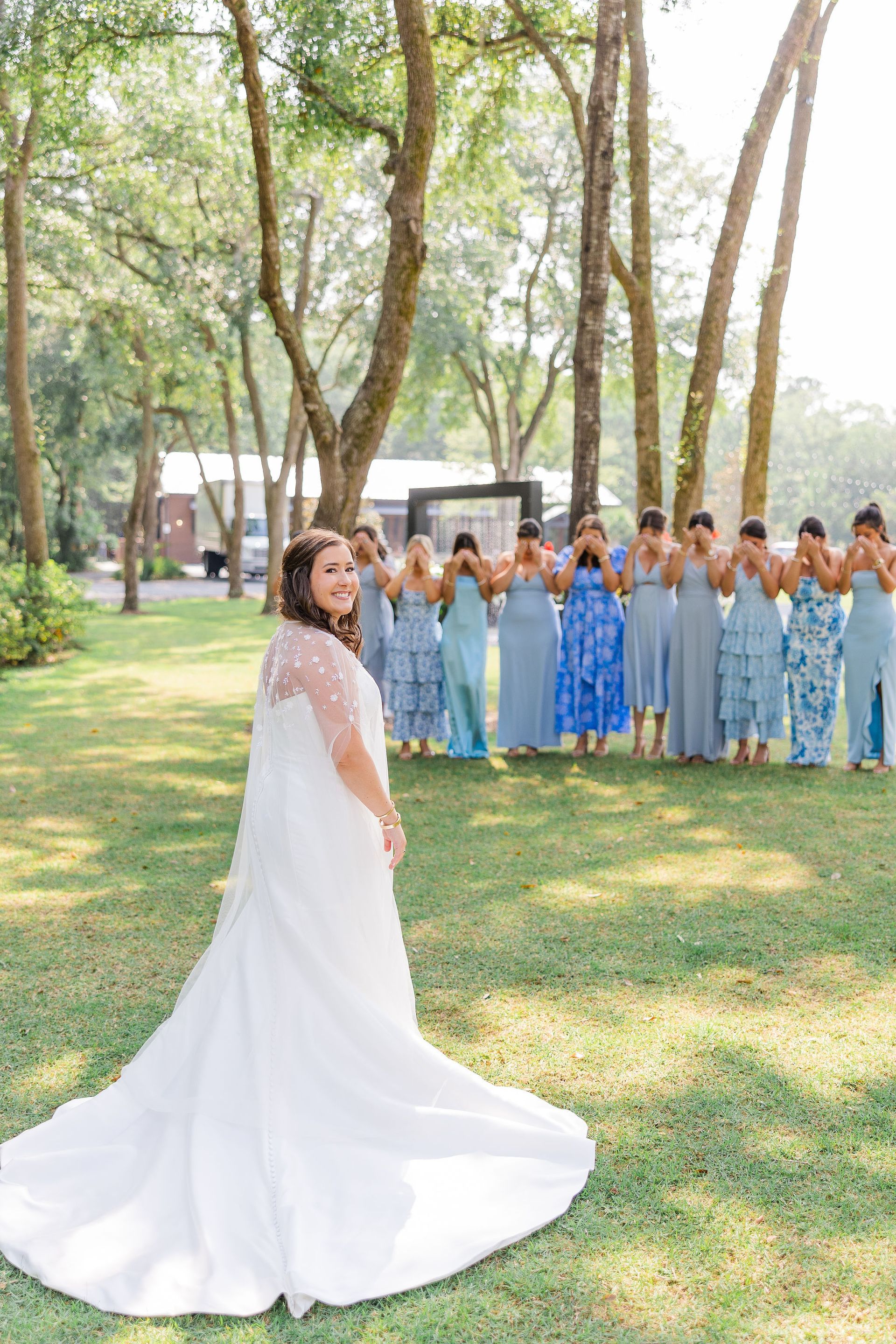 Bride in a white gown stands on a grassy lawn before a row of bridesmaids in blue dresses under tall trees.