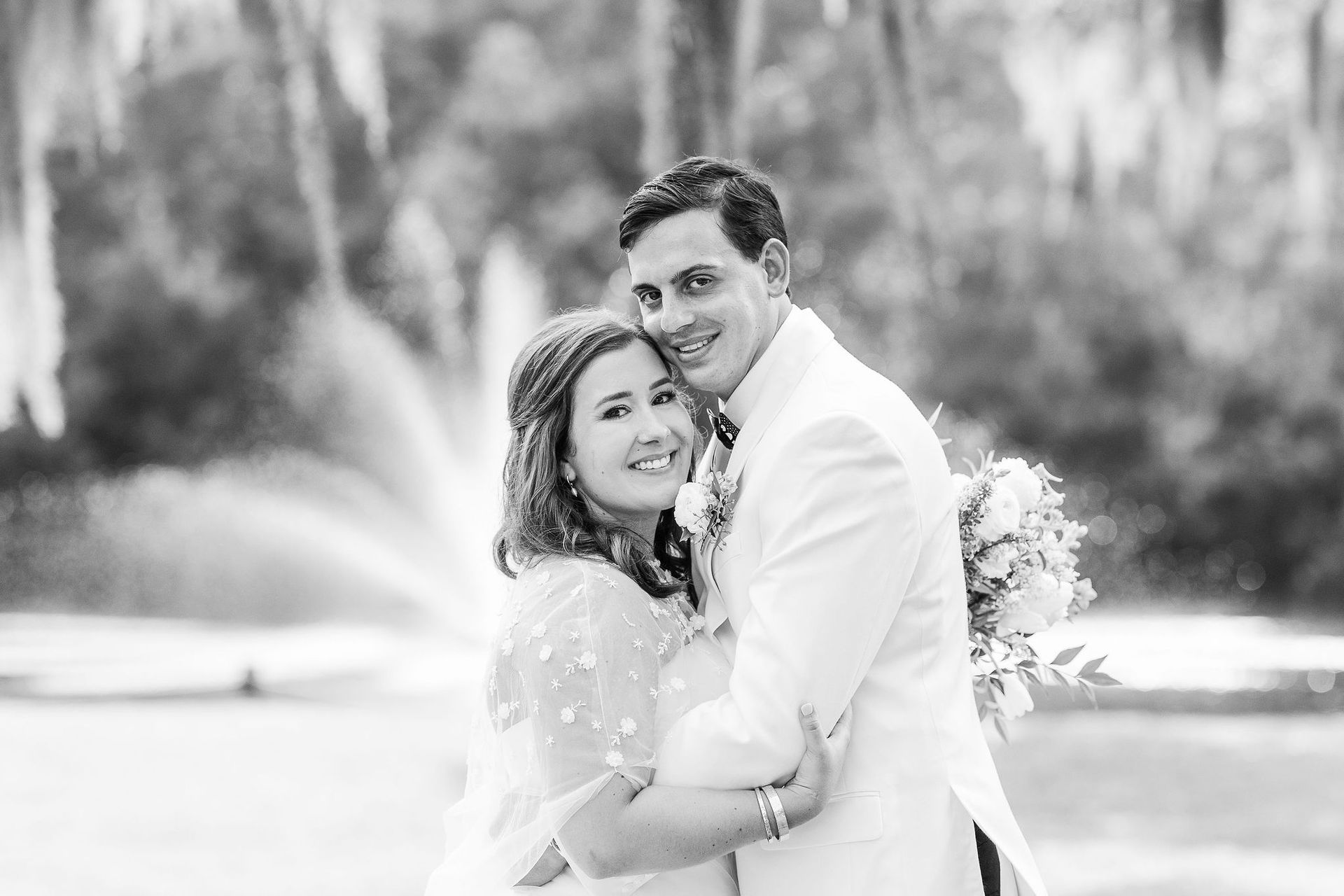 Smiling couple in white attire embracing outdoors near a fountain